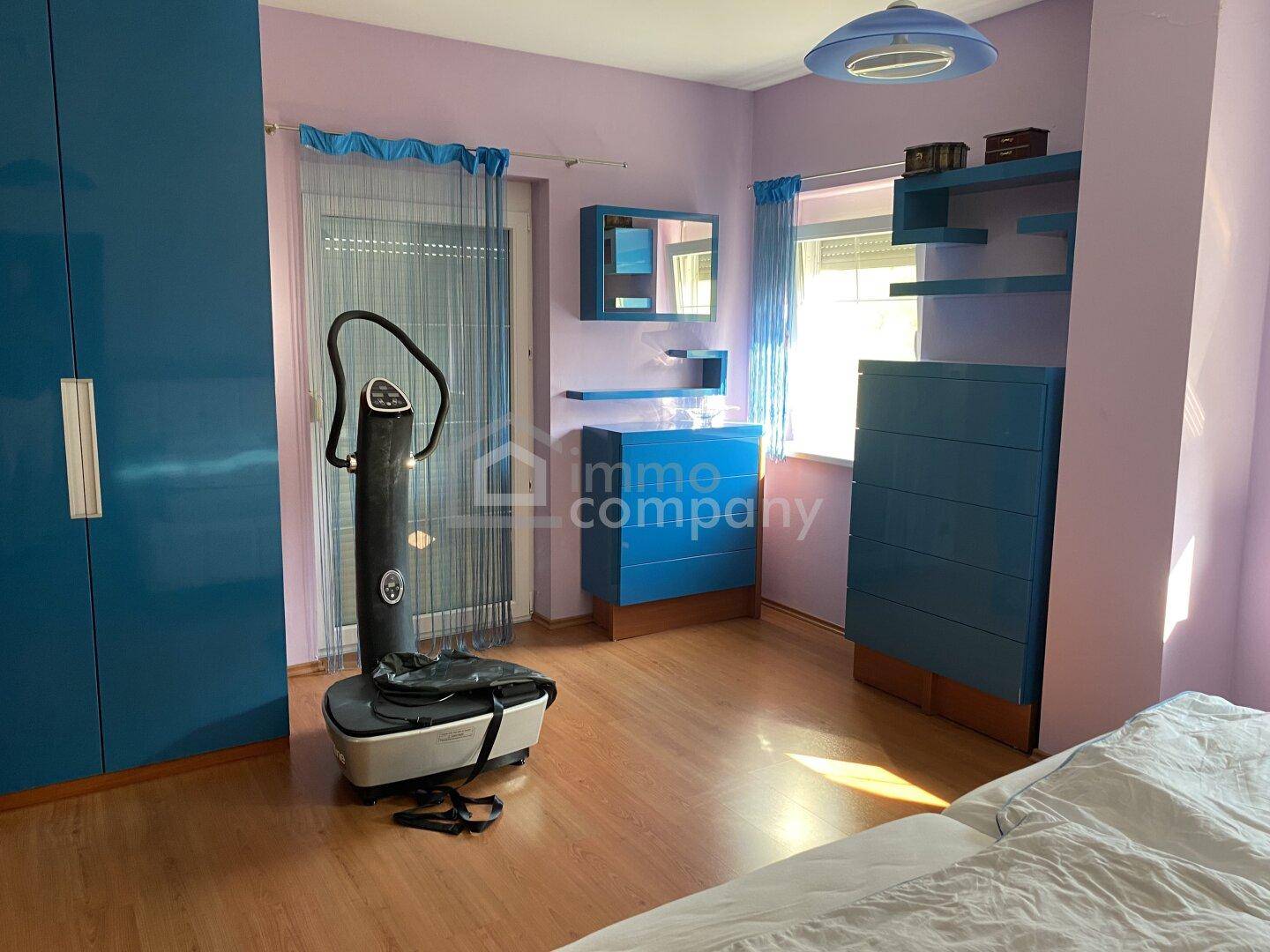 Schlafzimmer mit lila Wänden, blauen Möbeln und einem Fitnessgerät, hell und funktional.