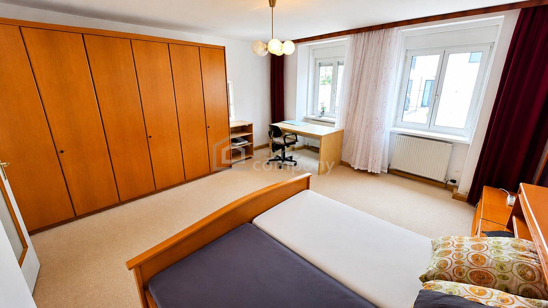 Schlafzimmer mit großem Holzkleiderschrank, Schreibtisch und zwei Fenstern für natürliches Licht.