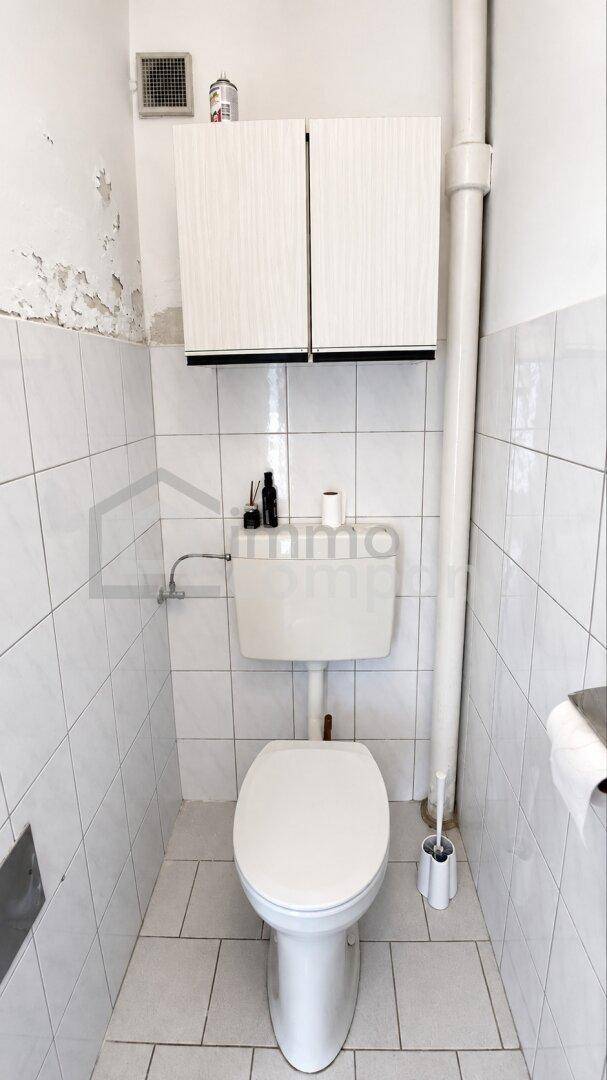 Kleines WC mit weiß gefliesten Wänden und Boden sowie einem Hängeschrank über der Toilette.