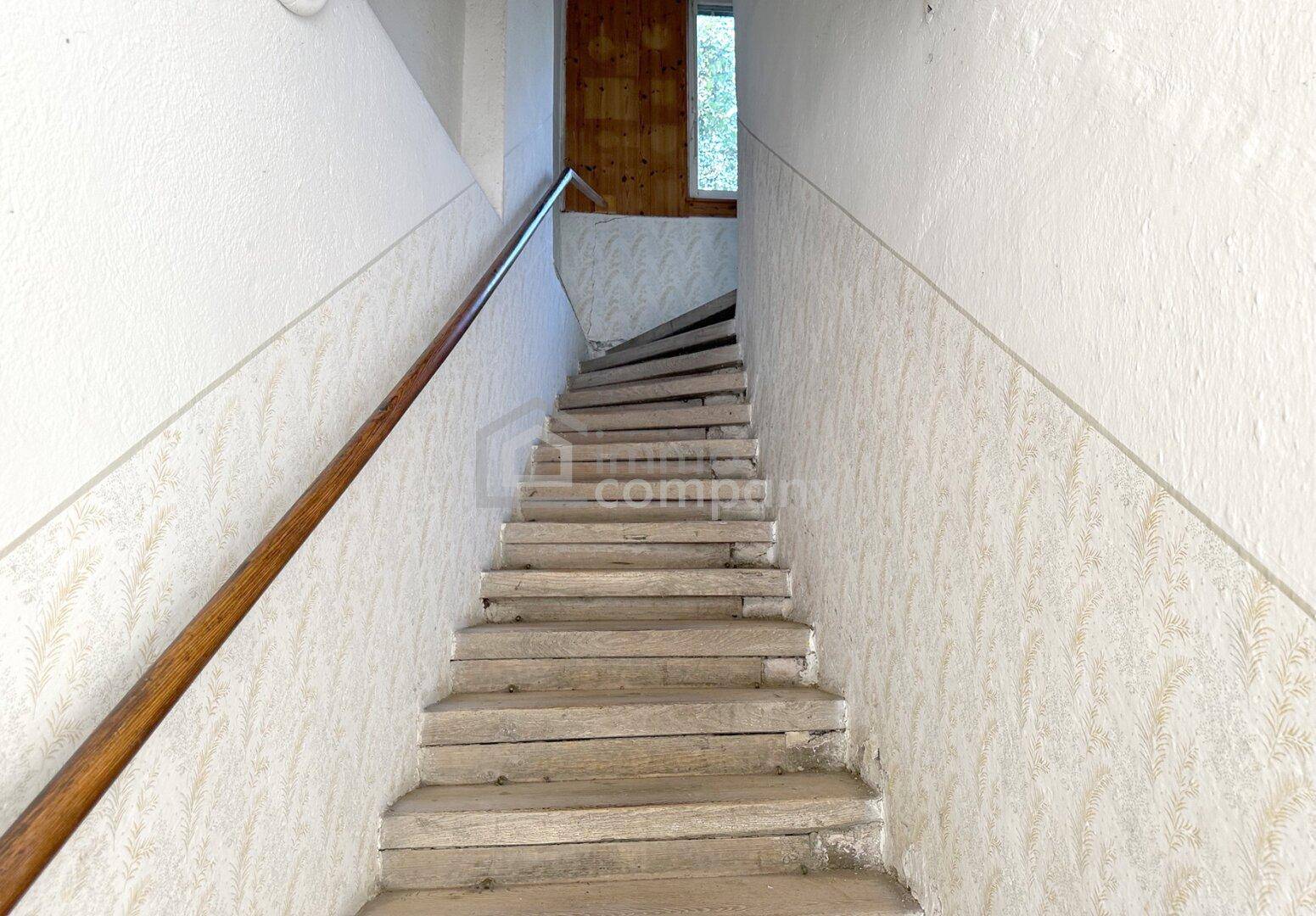 Treppenhaus mit Holzgeländer und abgenutzten Stufen, in renovierungsbedürftigem Zustand.