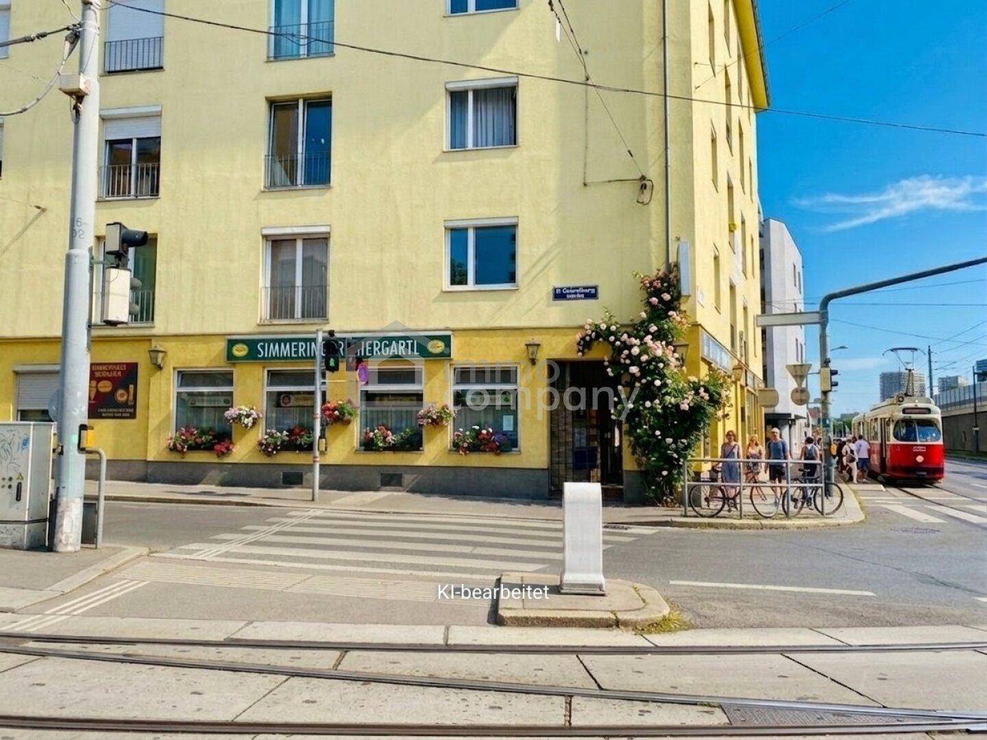 Gelbes Eckgebäude in urbaner Umgebung mit Balkonen, Fensterläden und einem Restaurant im Erdgeschoss, belebt durch eine Straßenbahn.
