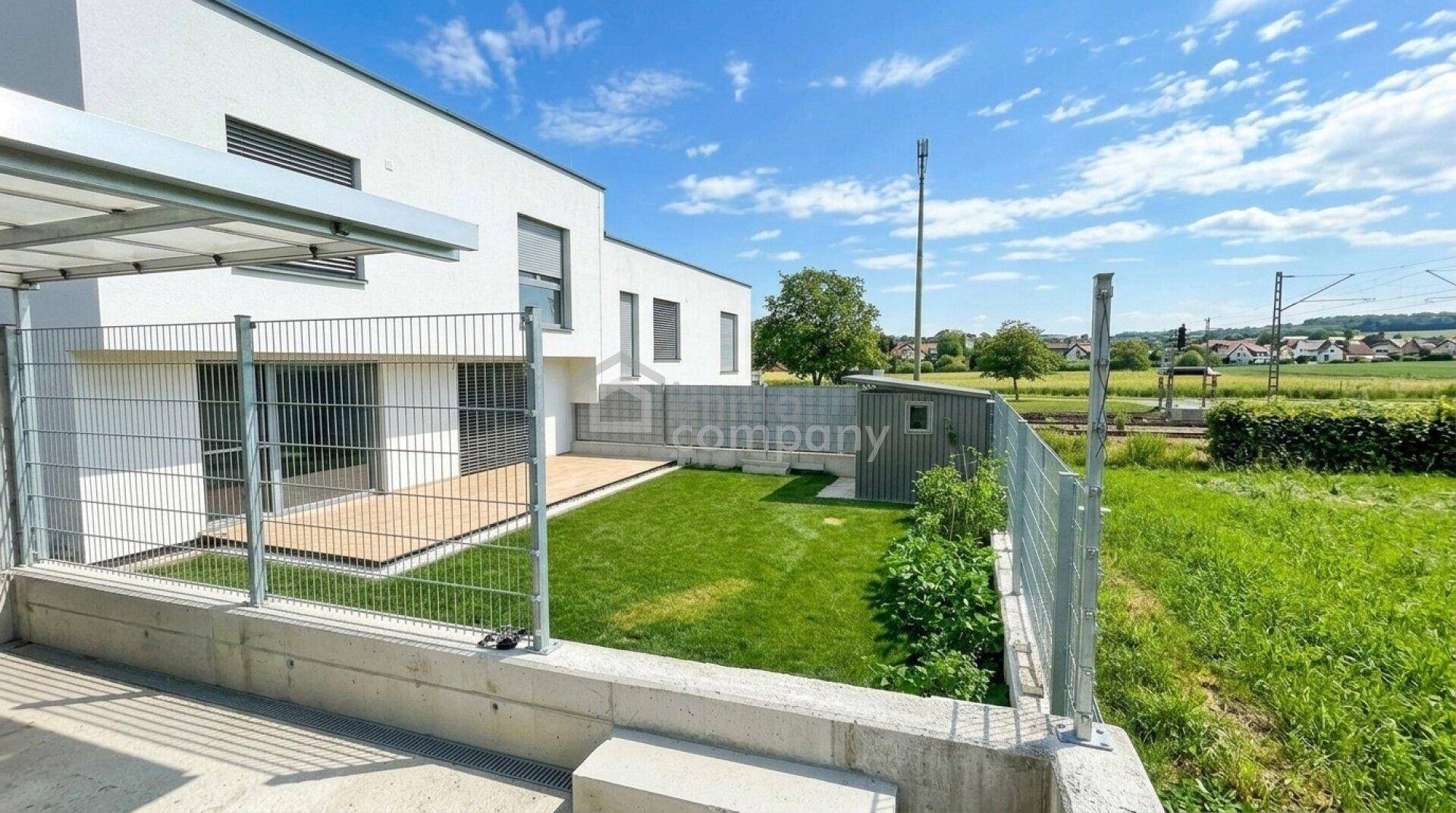 Modernes Wohngebäude mit Holzterrasse und Garten, angrenzend an grüne Felder und Bahngleise unter blauem Himmel.