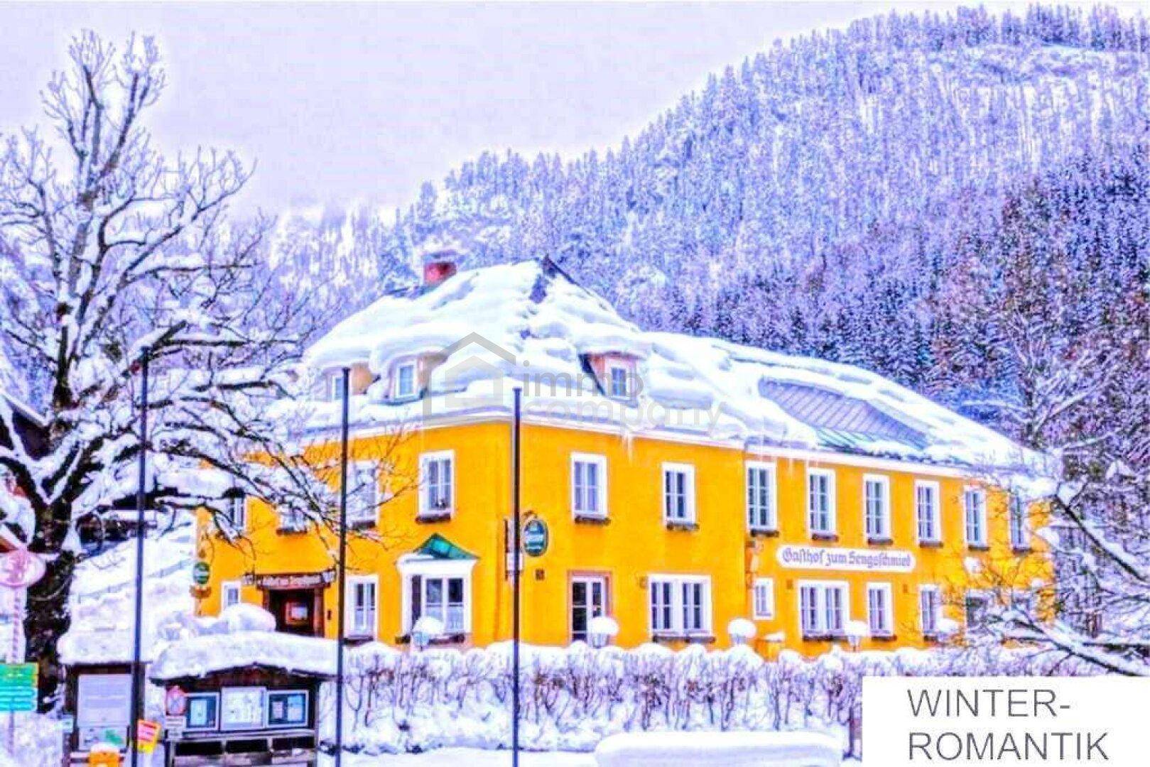 Winterliche Ansicht eines traditionellen Gasthofs mit schneebedecktem Dach und umliegender Landschaft.