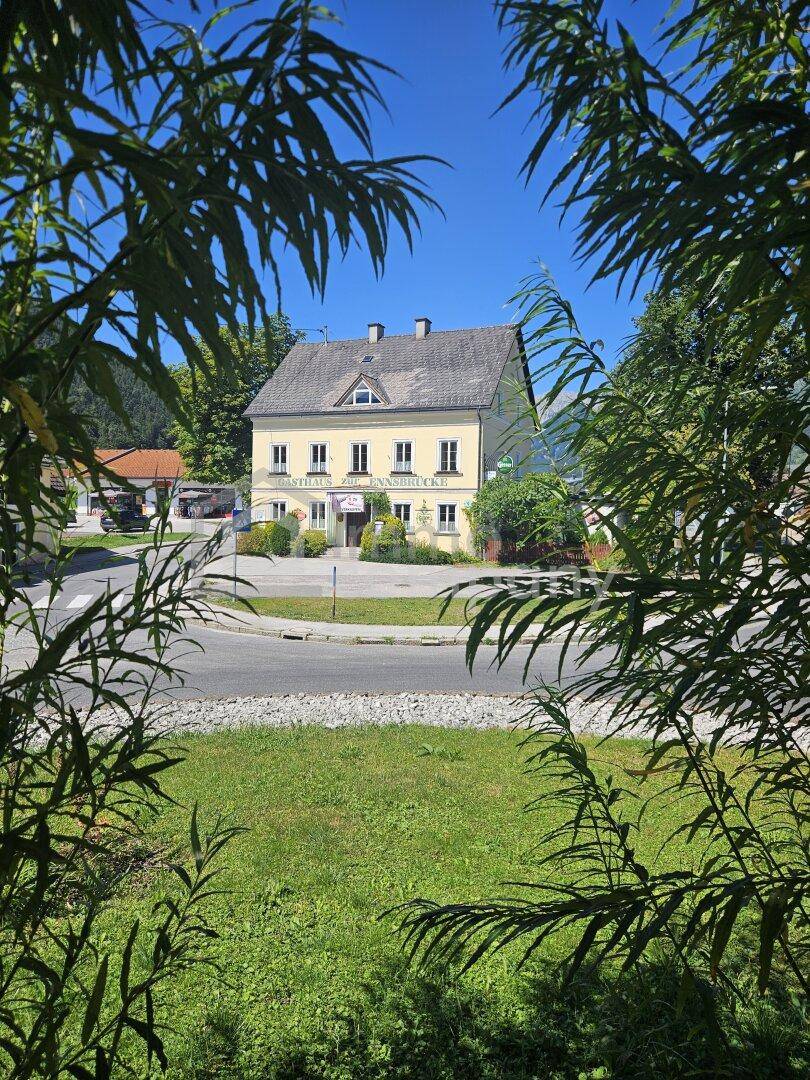 Das gelbe Gasthaus ist durch grüne Blätter eingerahmt, mit einem Kreisverkehr und Bergen im Hintergrund.