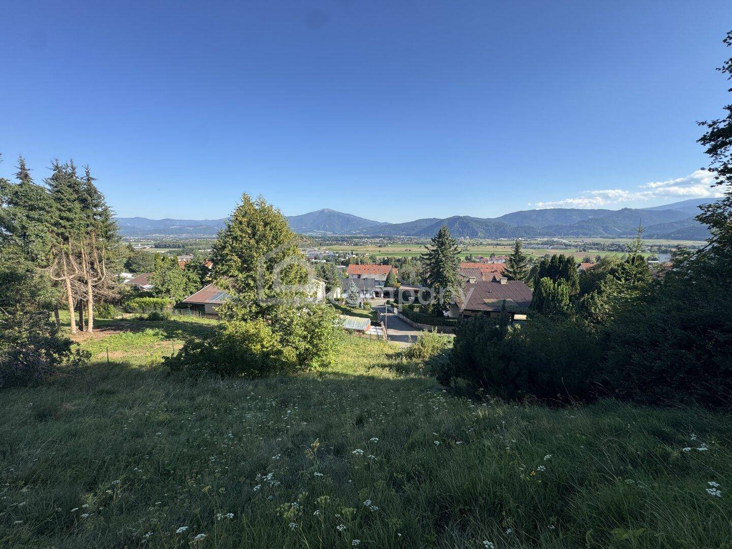 Panoramablick über eine grüne Hügellandschaft mit Häusern und Bergen am Horizont unter blauem Himmel.