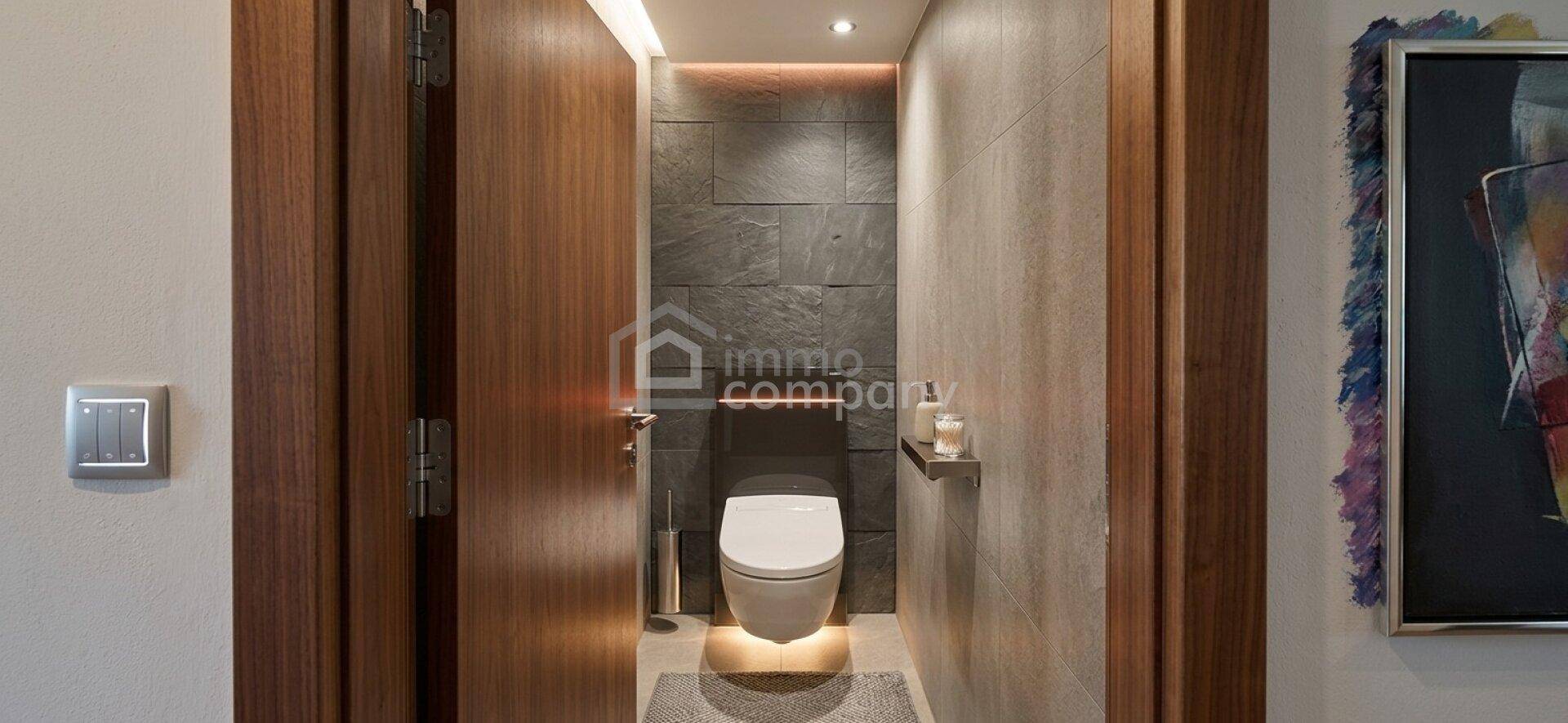 Modernes Gäste-WC mit Wand-WC, dunklen Fliesen und indirekter Beleuchtung.