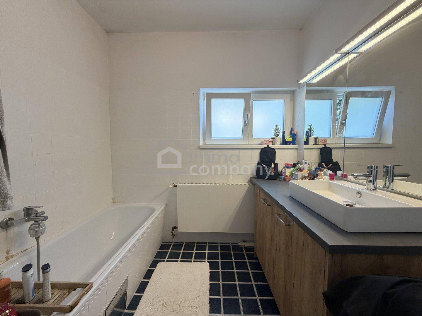Badezimmer mit Badewanne, Einzelwaschbecken, Fenster und dunklen Fliesen auf dem Boden.
