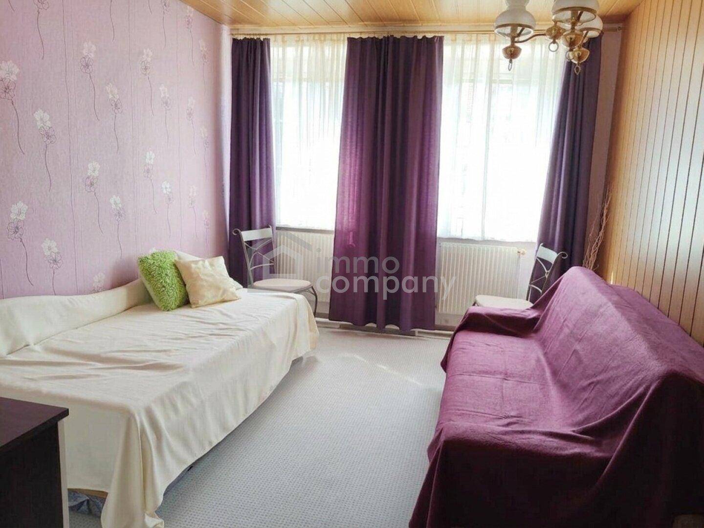 Schlafzimmer mit zwei Betten, lila Vorhängen und floraler Tapete, hell und freundlich gestaltet.