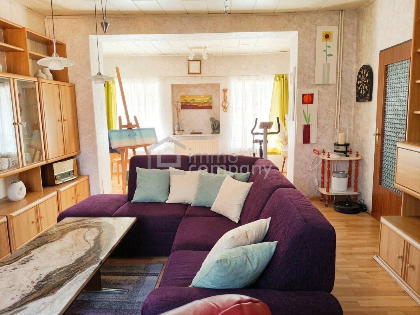 Geräumiges Wohnzimmer mit violetter Eckcouch, Holzboden und offenem Übergang zu einem weiteren Raum.
