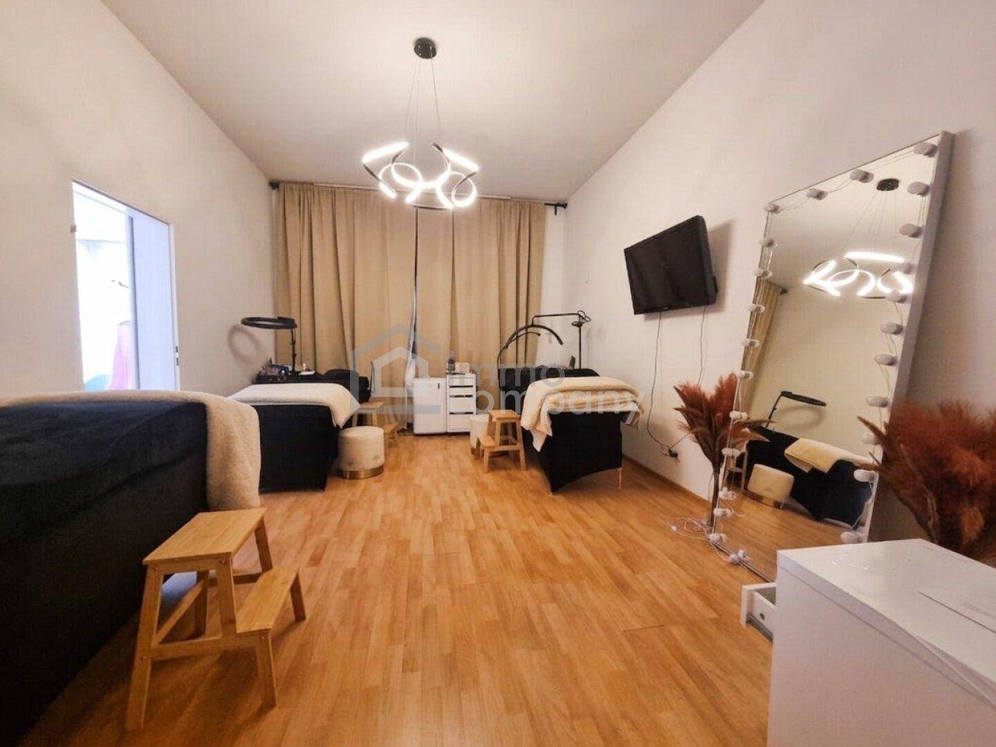 Geräumiger Behandlungsraum mit Holzboden, zwei Liegen, einem großen beleuchteten Spiegel und moderner Deckenleuchte.