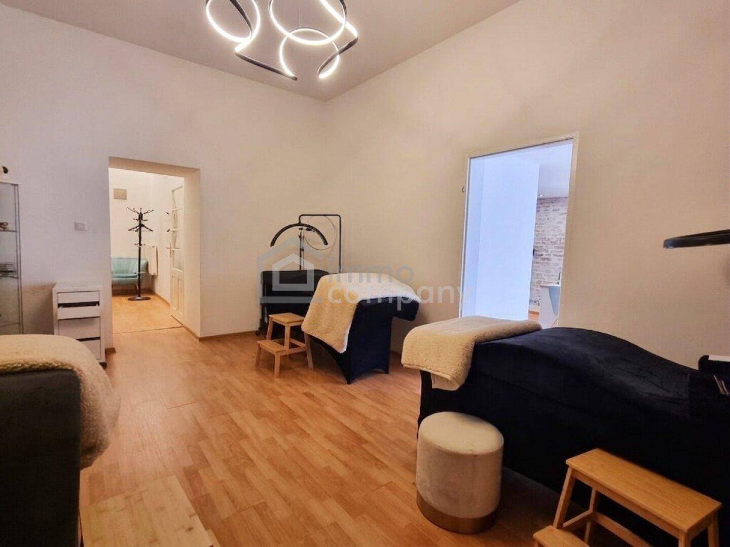 Behandlungsraum mit Holzboden und zwei Liegen, Blick auf einen angrenzenden Flur und moderne Deckenleuchte.