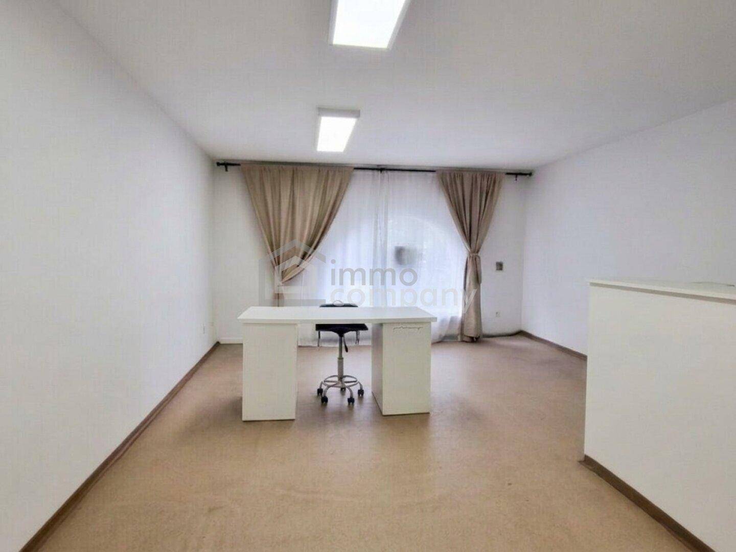 Leerer Büroraum mit einem Schreibtisch, Bürostuhl und großen Fenstern mit beigen Vorhängen.