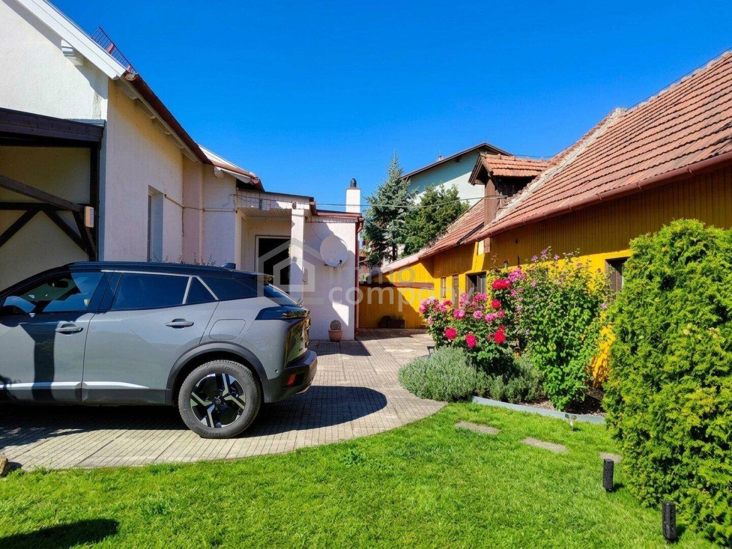 Gepflegter Gartenbereich mit Rasenfläche, gepflastertem Weg, blühenden Rosen und Parkplatz.