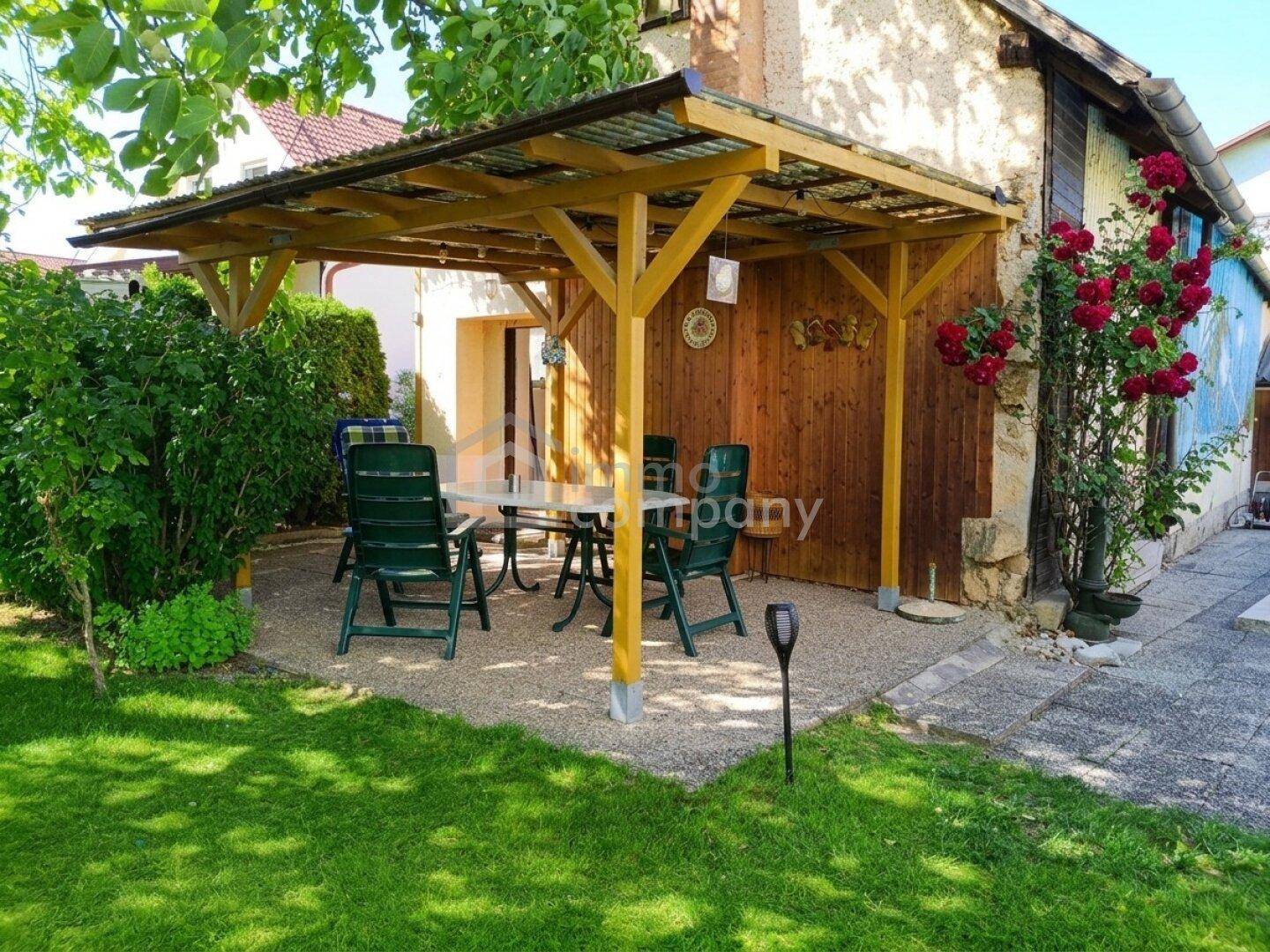 Überdachter Sitzbereich im Garten mit Holzdach und Gartenmöbeln, umgeben von Grünflächen.