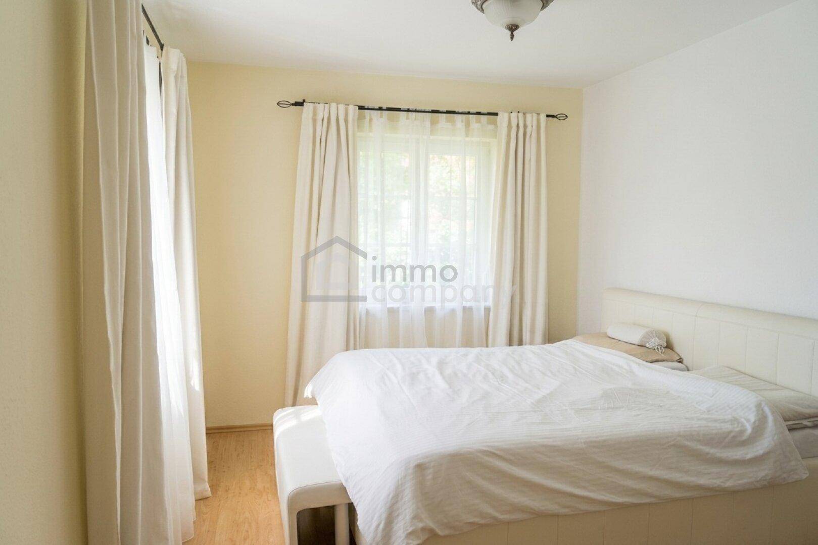 Gemütliches Schlafzimmer mit Doppelbett, hellem Laminatboden und Fenster mit Vorhängen.
