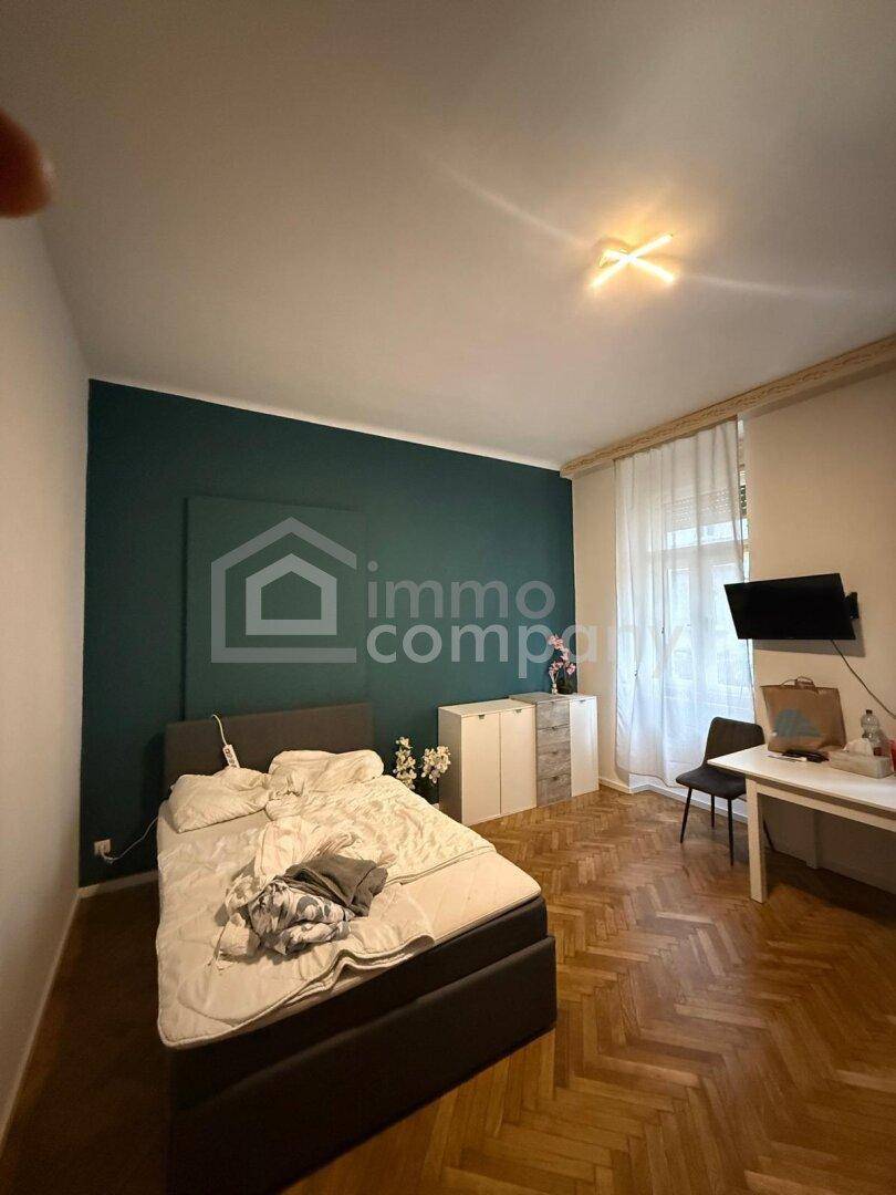 Helles Schlafzimmer mit Parkettboden, großem Fenster, Schreibtisch und Wandfernseher.