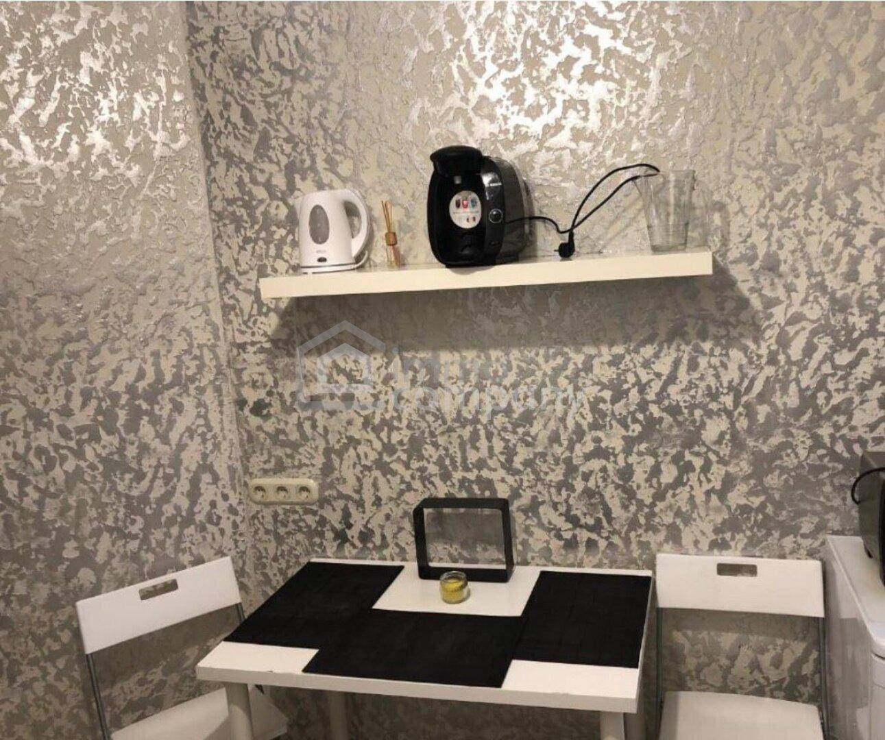 Kleine Küchenzeile mit Esstisch, Wasserkocher und Kaffeemaschine vor strukturierter Tapete.