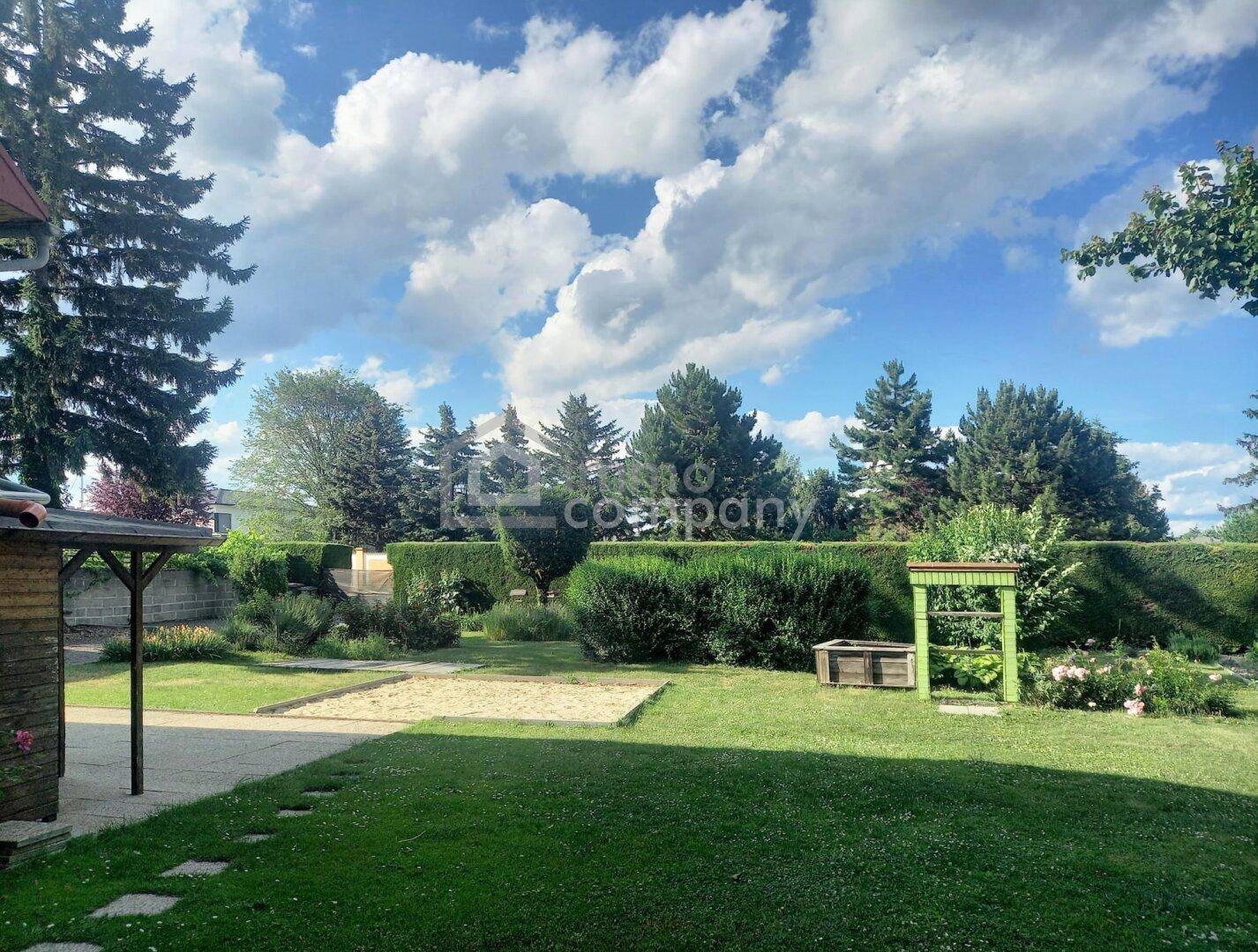 Großer Garten mit Rasenfläche, Sandkasten und hohen Bäumen unter blauem Himmel.