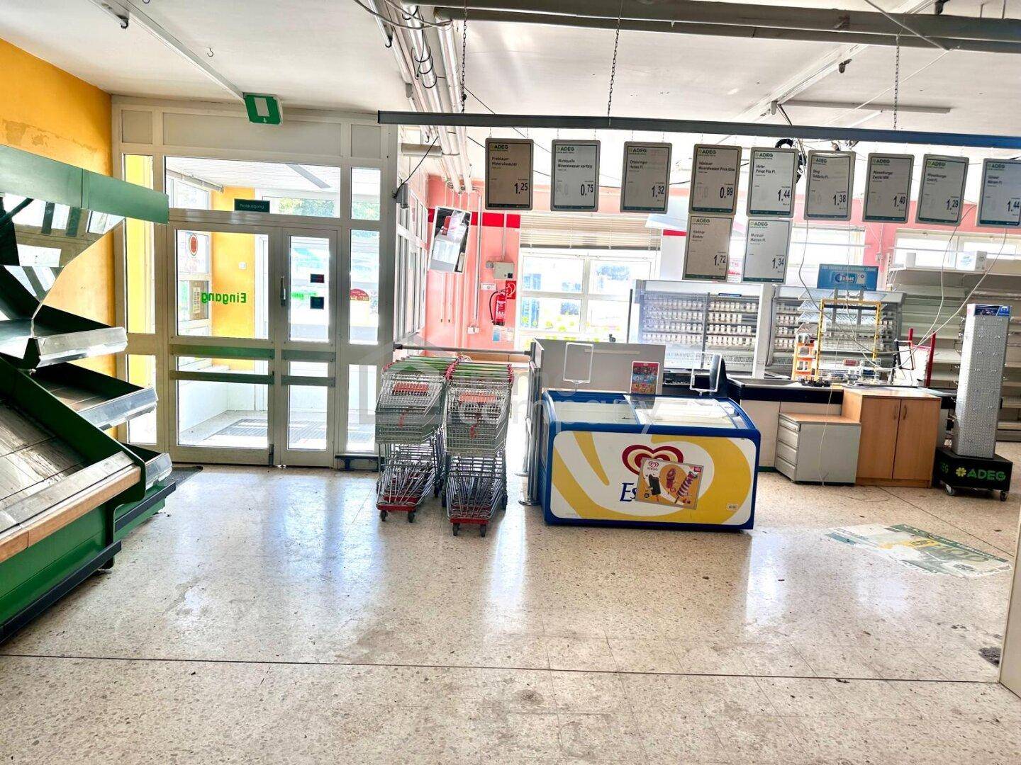 Eingangsbereich eines Geschäfts mit Glastüren, Einkaufswagen und einer Gefriertruhe, gut beleuchtet.