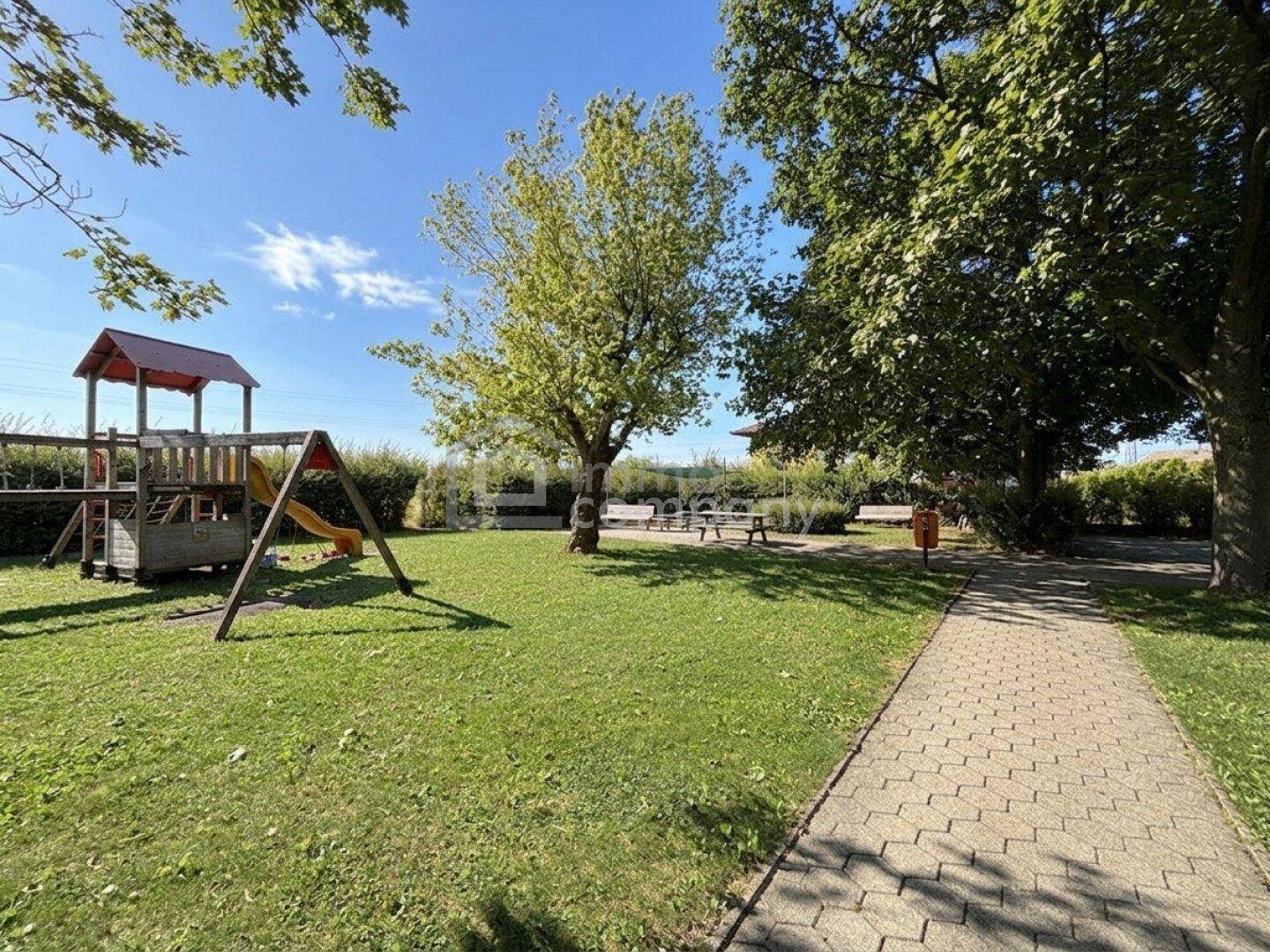Grüner Gartenbereich mit Kinderspielplatz, Schaukel und Rutsche unter schattenspendenden Bäumen.