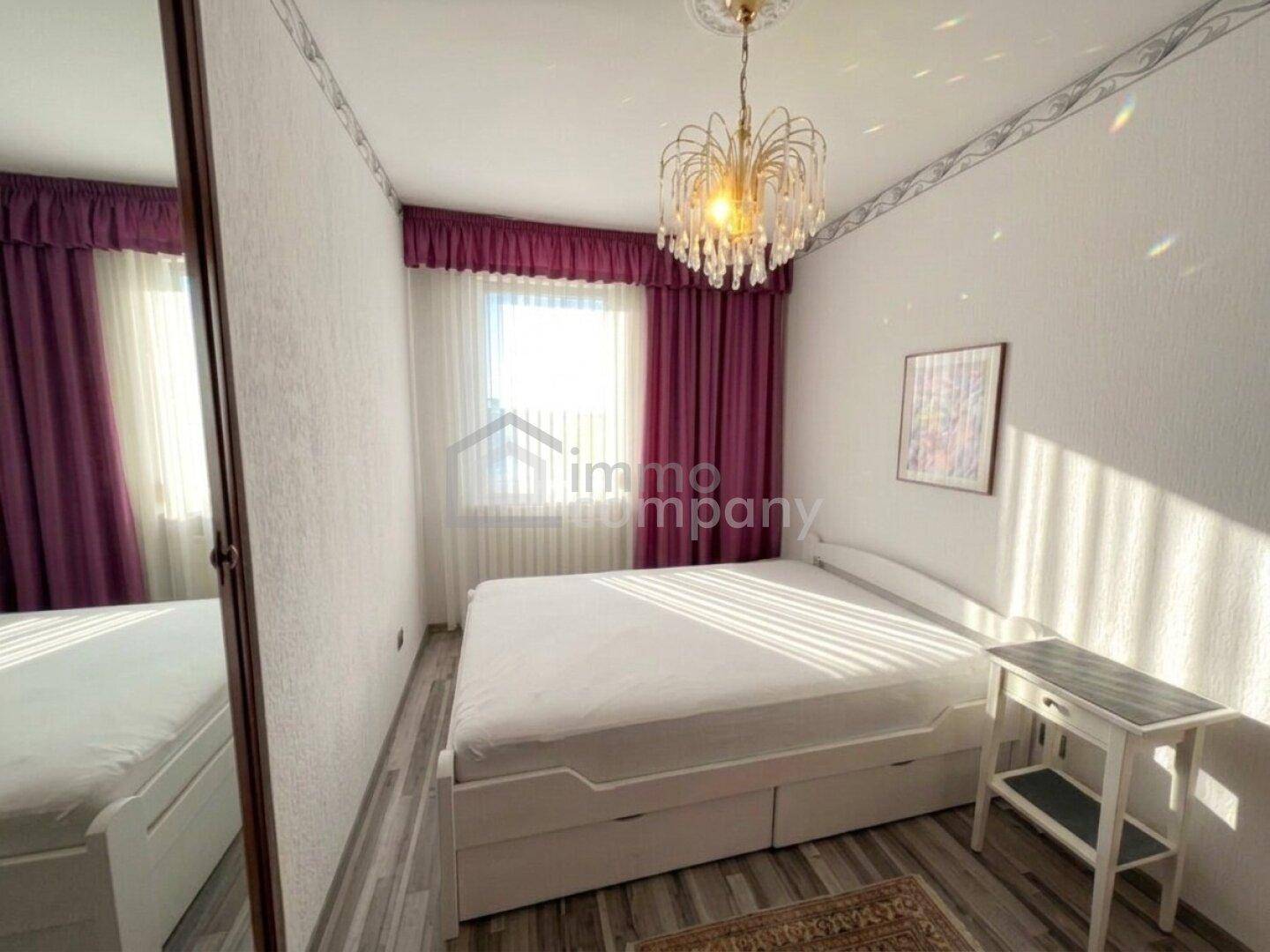 Gemütliches Schlafzimmer mit Doppelbett, lila Vorhängen und einem Kronleuchter, hell durch Tageslicht.