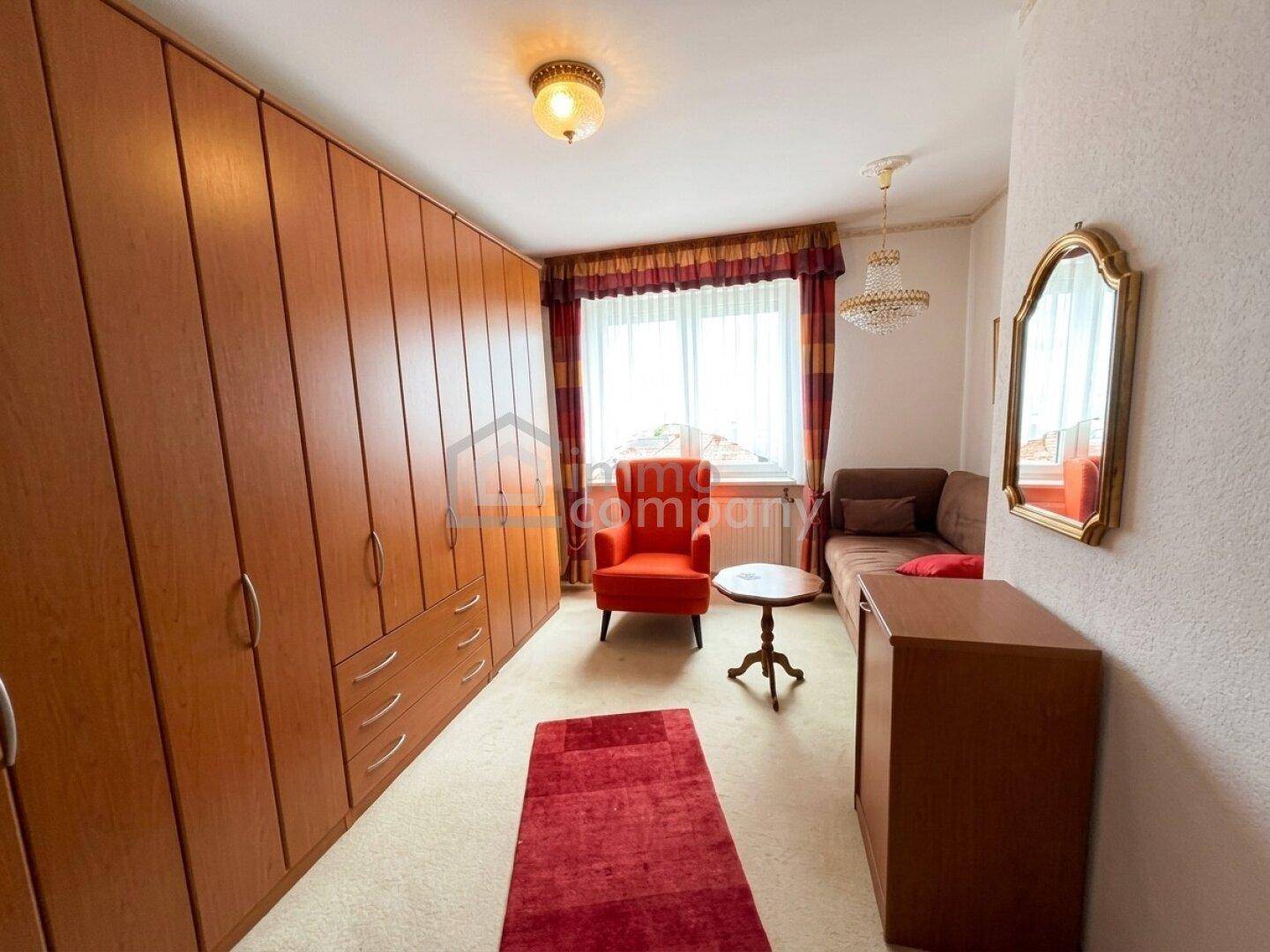Zimmer mit großen Einbauschränken, rotem Sessel und Sofa, hell durch das Fenster.