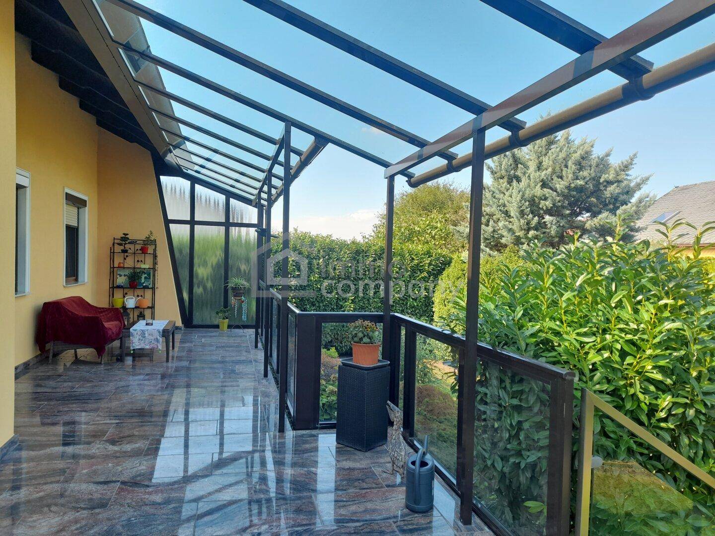Überdachte Terrasse mit Fliesenboden, Glasgeländer und Blick ins Grüne.