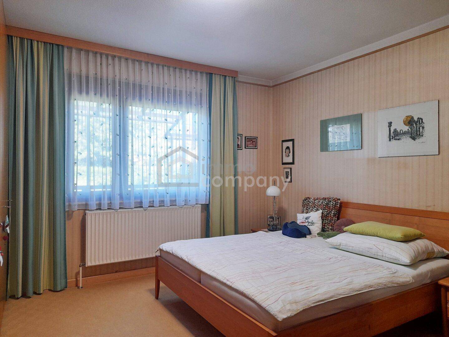 Schlafzimmer mit Doppelbett, gemusterter Tapete und Fenster mit Vorhängen, bietet viel Tageslicht.