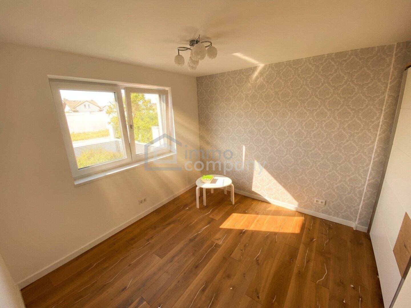 Helles Schlafzimmer mit Holzboden, Fenster und dezenter Tapete, gut beleuchtet.