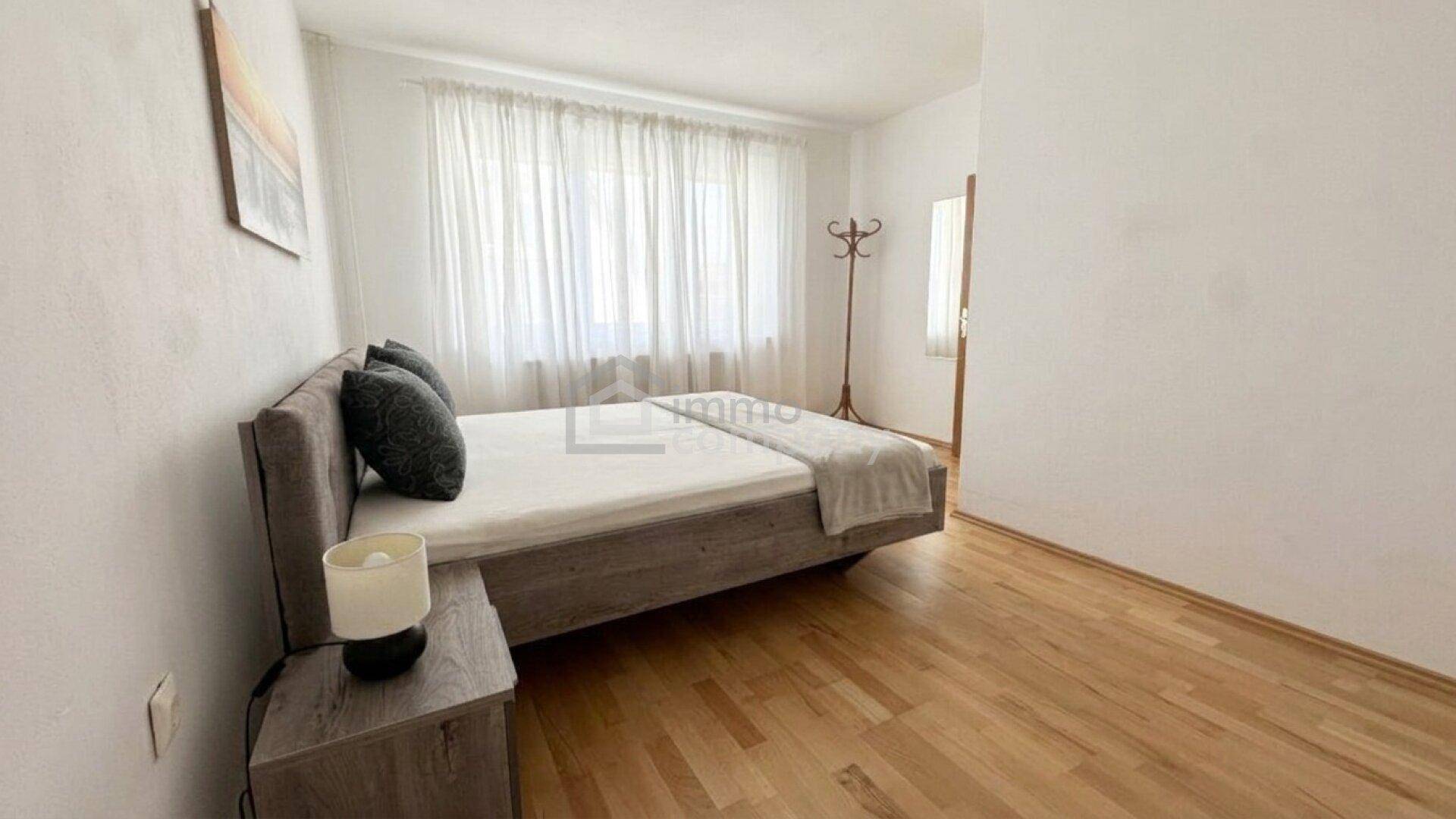 Schlafzimmer mit modernem Bett, Nachttisch und hellem Laminatboden, gut beleuchtet und schlicht eingerichtet.