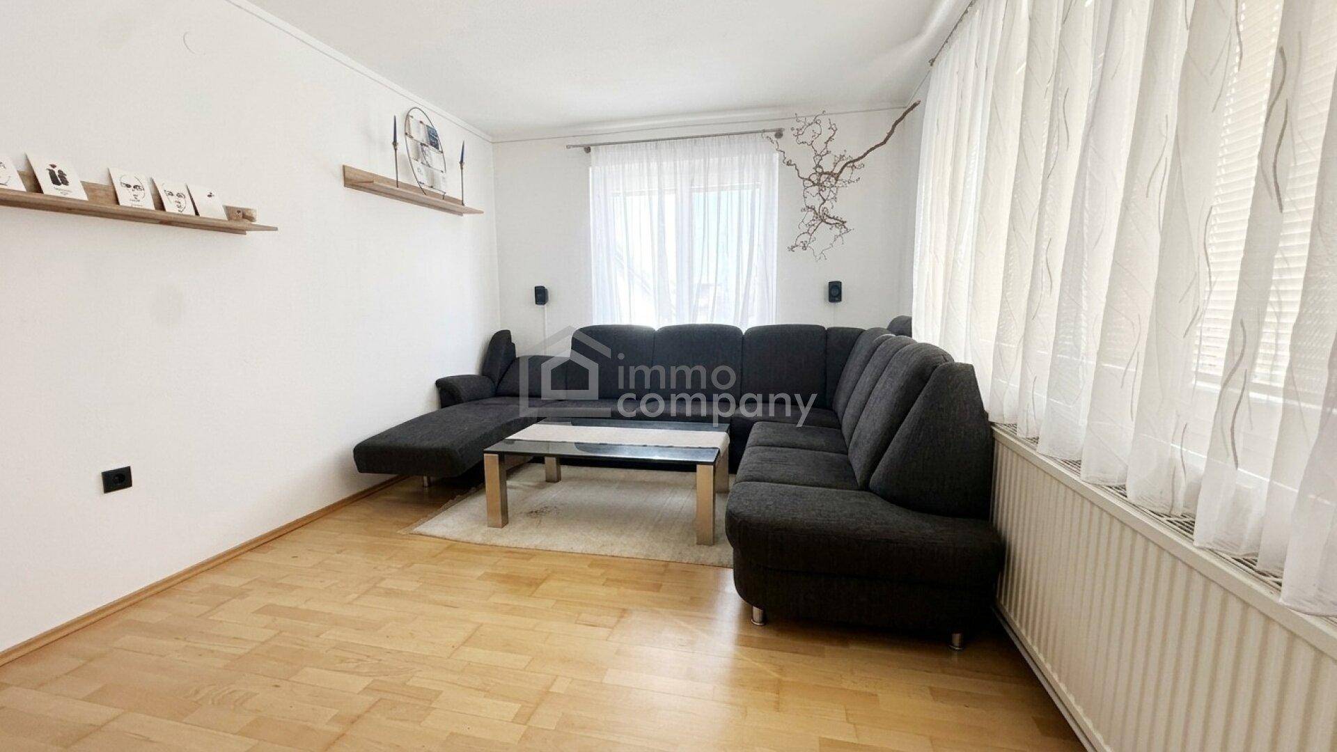 Gemütliches Wohnzimmer mit dunklem Ecksofa, Couchtisch und hellem Laminatboden, gut beleuchtet.