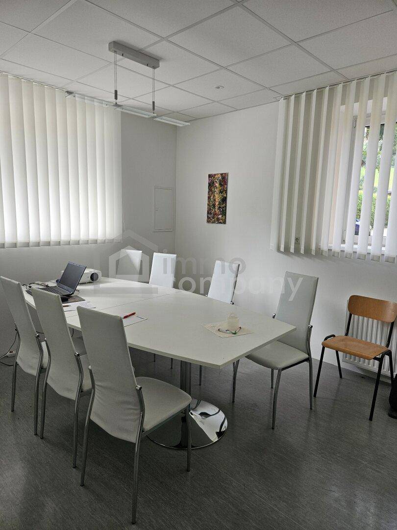 Konferenzraum mit großem Tisch, weißen Stühlen und vertikalen Jalousien an den Fenstern.