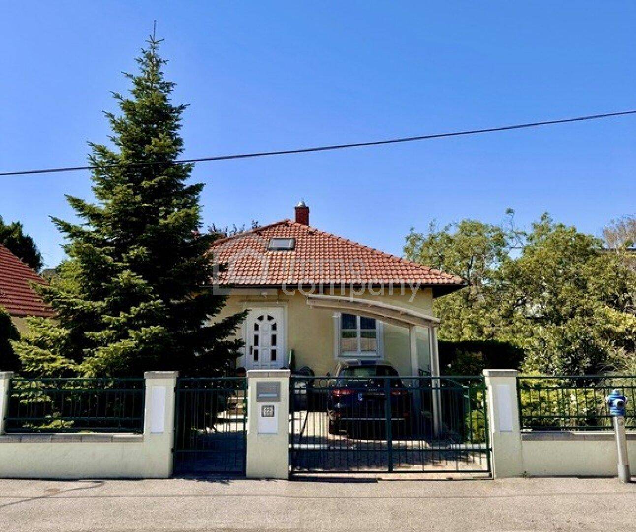 Vorderansicht eines gelben Hauses mit rotem Ziegeldach und einem grünen Gartentor.