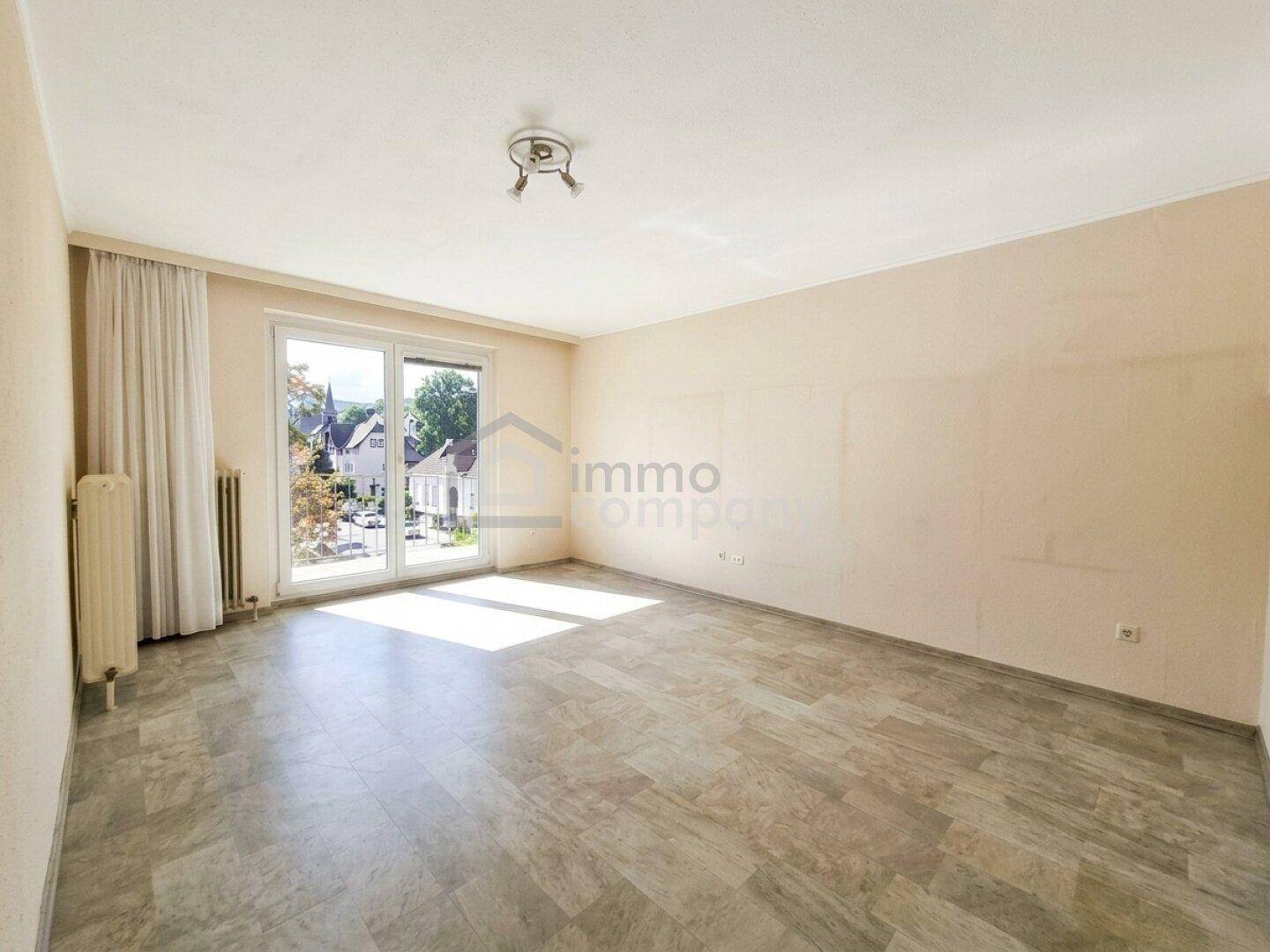 Heller, leerer Wohnraum mit großem Fenster und Zugang zum Außenbereich, ausgestattet mit Heizkörper.