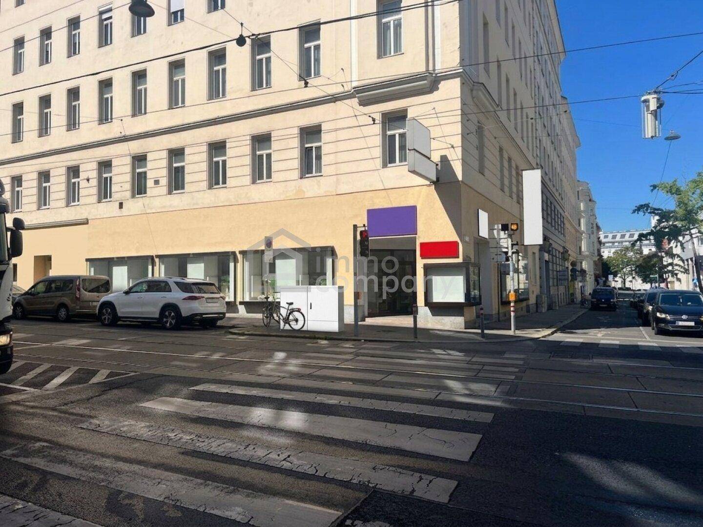 Eckgebäude an einer belebten Kreuzung mit Schaufenstern im Erdgeschoss und Straßenbahnschienen.