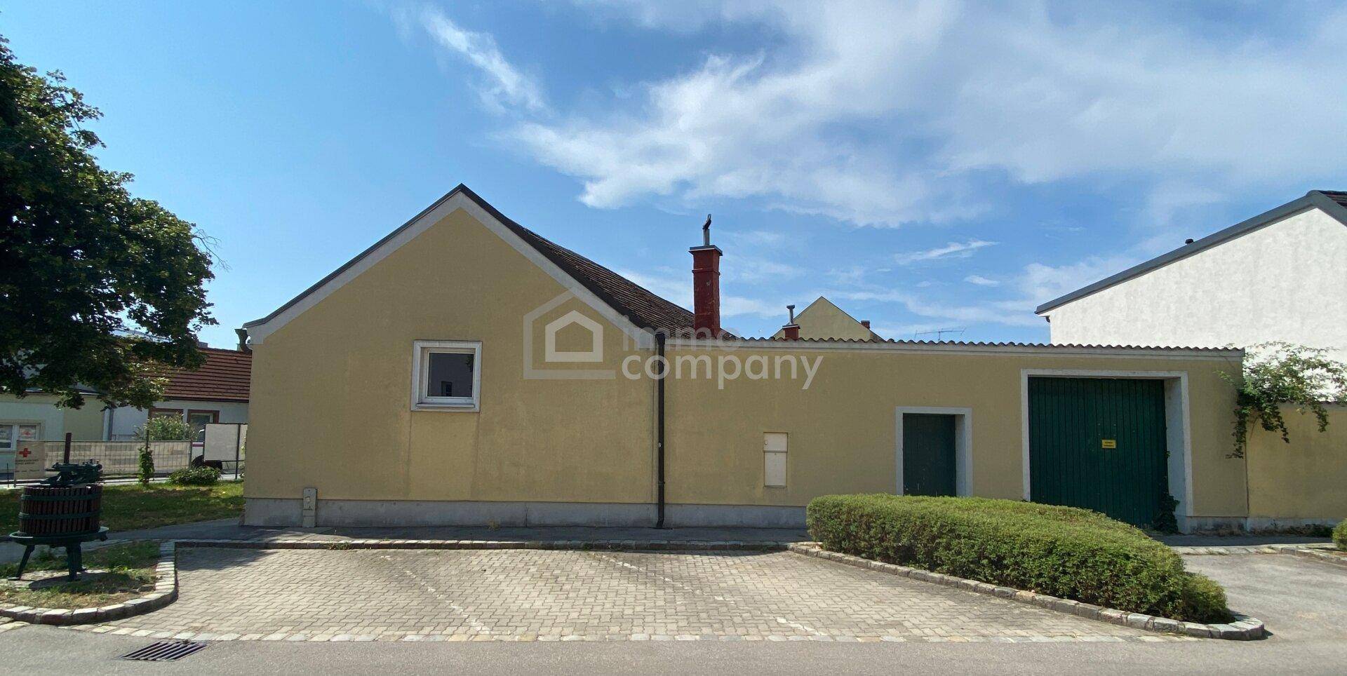 Rückansicht eines gelben Gebäudes mit Garage, gepflastertem Bereich und Hecke.