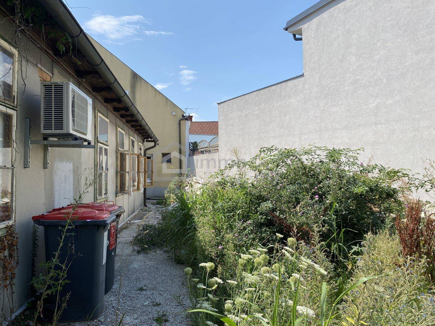 Seitenansicht eines Gebäudes mit Klimaanlage, alten Fenstern und einem verwilderten Gartenbereich.