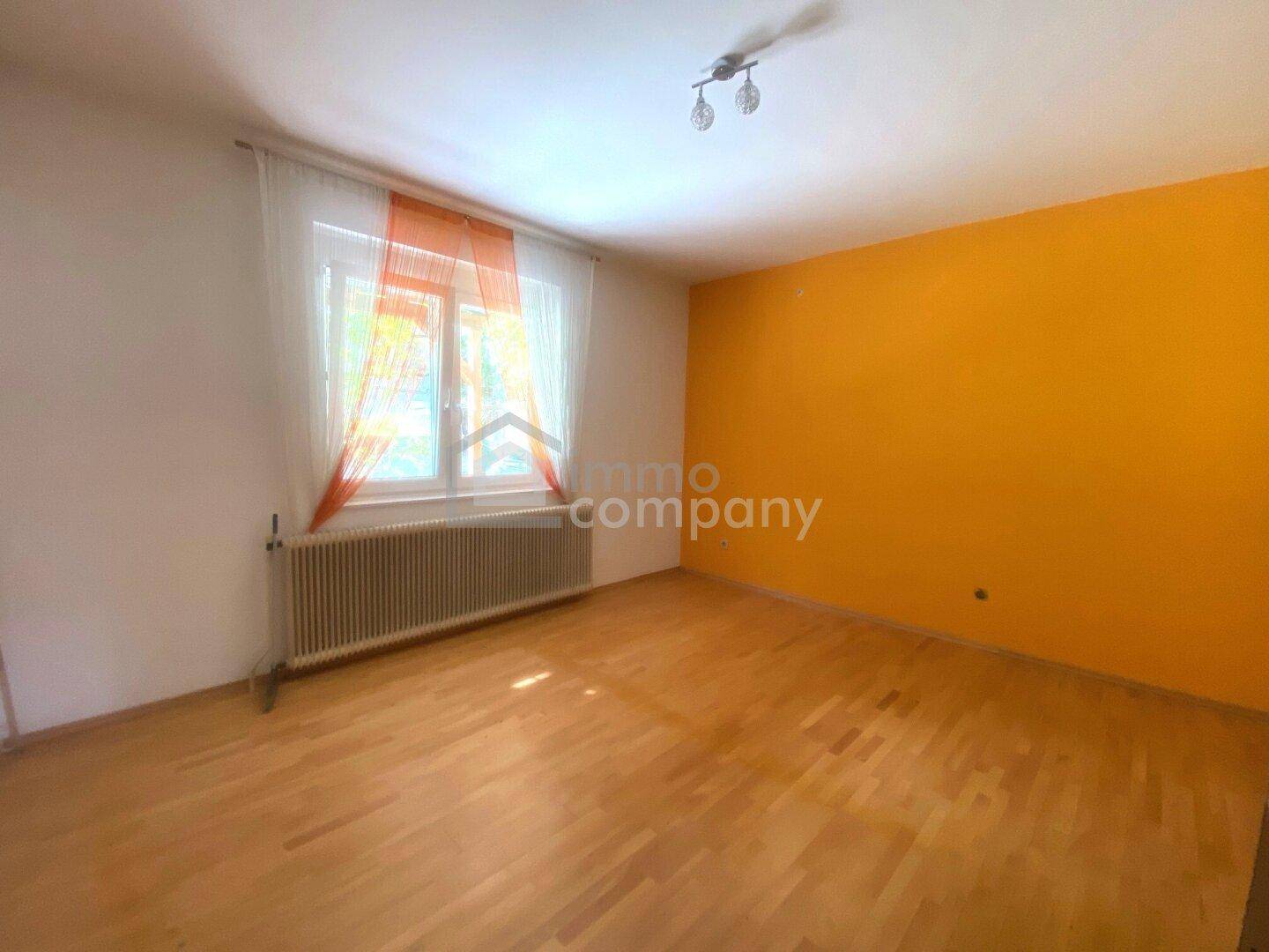 Helles Zimmer mit Holzboden, Fenster und einer auffälligen orangefarbenen Wand.