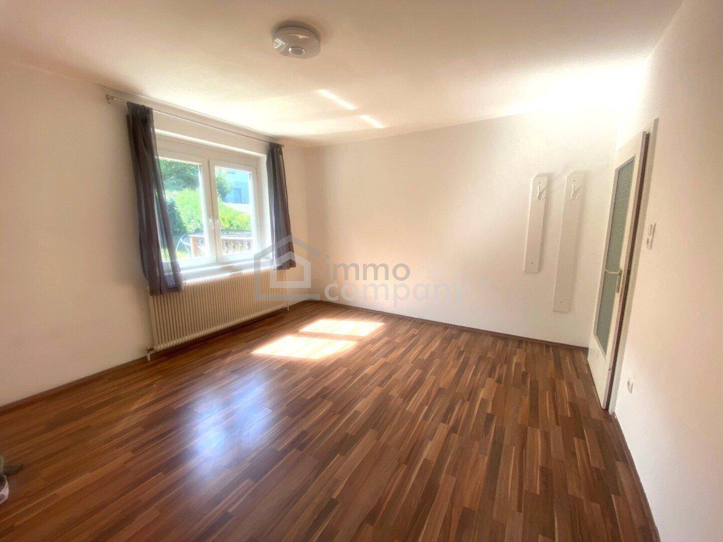 Helles Zimmer mit dunklem Holzboden und einem Fenster mit Blick ins Grüne.