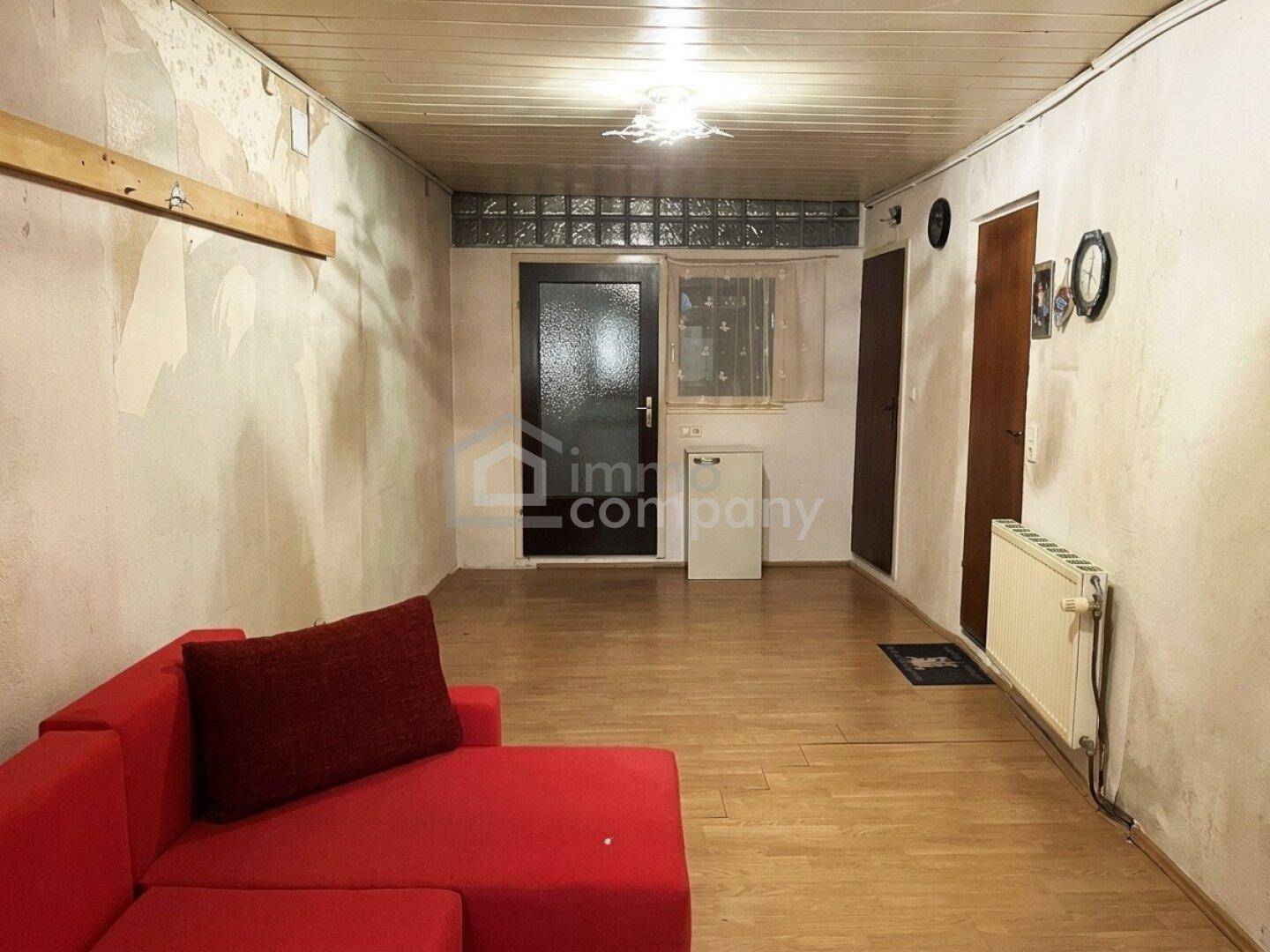 Wohnraum mit rotem Sofa, Holzfußboden und Renovierungsbedarf an den Wänden.
