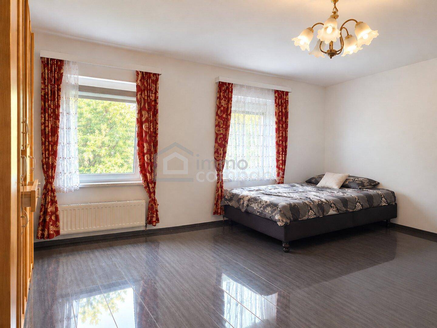 Schlafzimmer mit dunklem Laminatboden, Doppelbett und großen Fenstern mit rotem Vorhang.