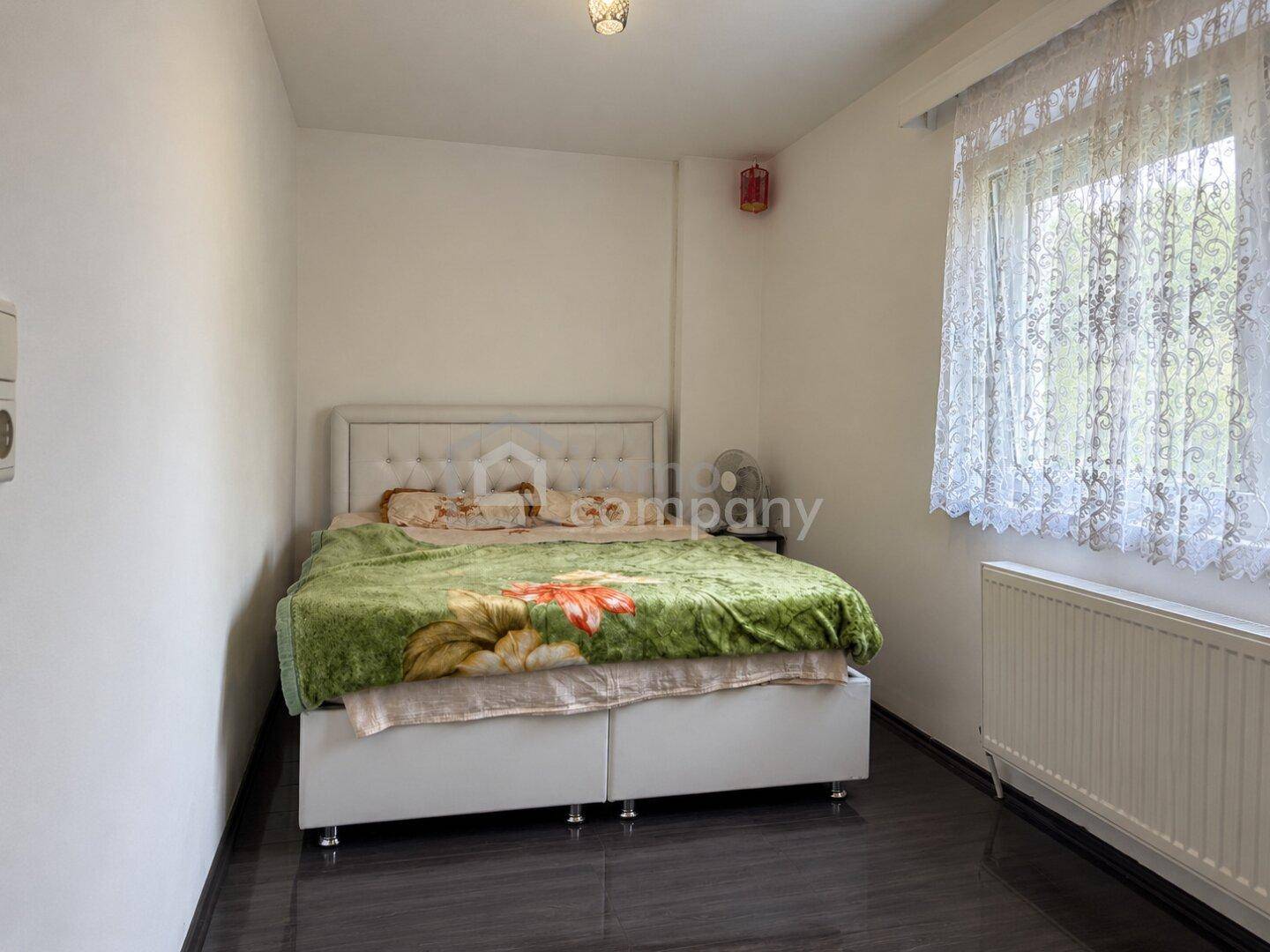 Helles Schlafzimmer mit Doppelbett, weißem Kopfteil und Fenster mit Spitzenvorhang.