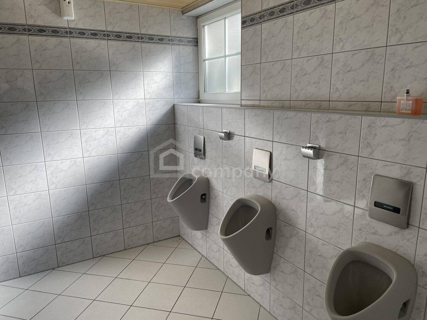 Herren-WC mit mehreren Urinalen und hell gefliesten Wänden, ausgestattet mit einem Fenster.