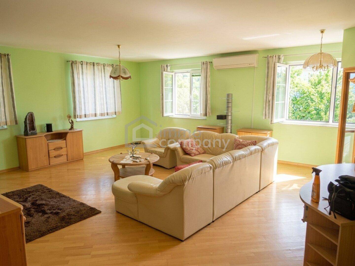 Geräumiges Wohnzimmer mit hellgrünen Wänden, beigem Sofa, Holzboden und Klimaanlage.