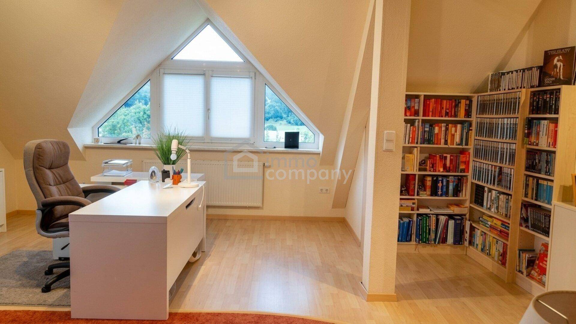 Helles Dachgeschoss-Büro mit schrägen Wänden, großem Fenster und Holzboden.