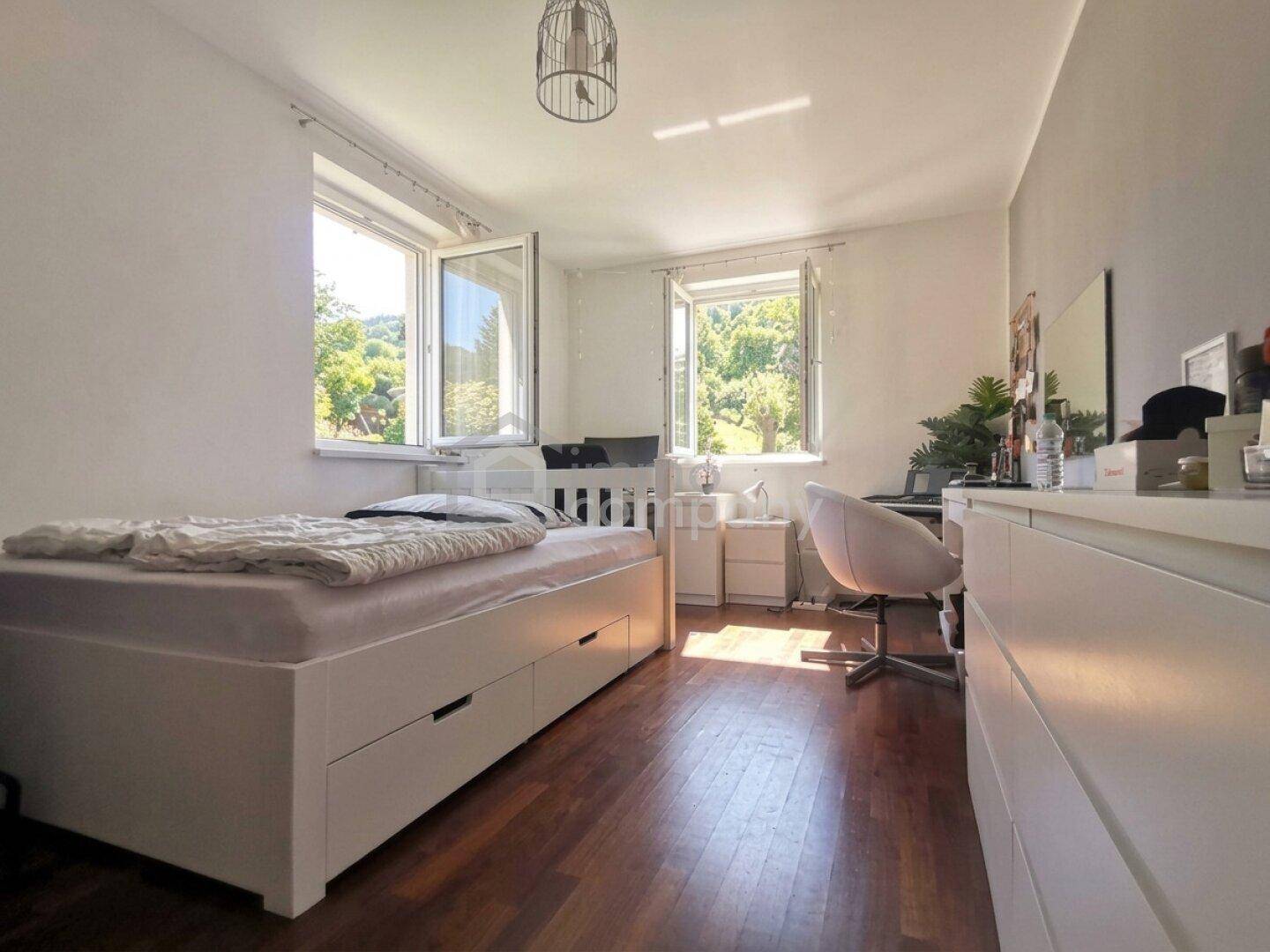 Helles Schlafzimmer mit Einzelbett, Schreibtisch und Blick ins Grüne durch zwei Fenster.