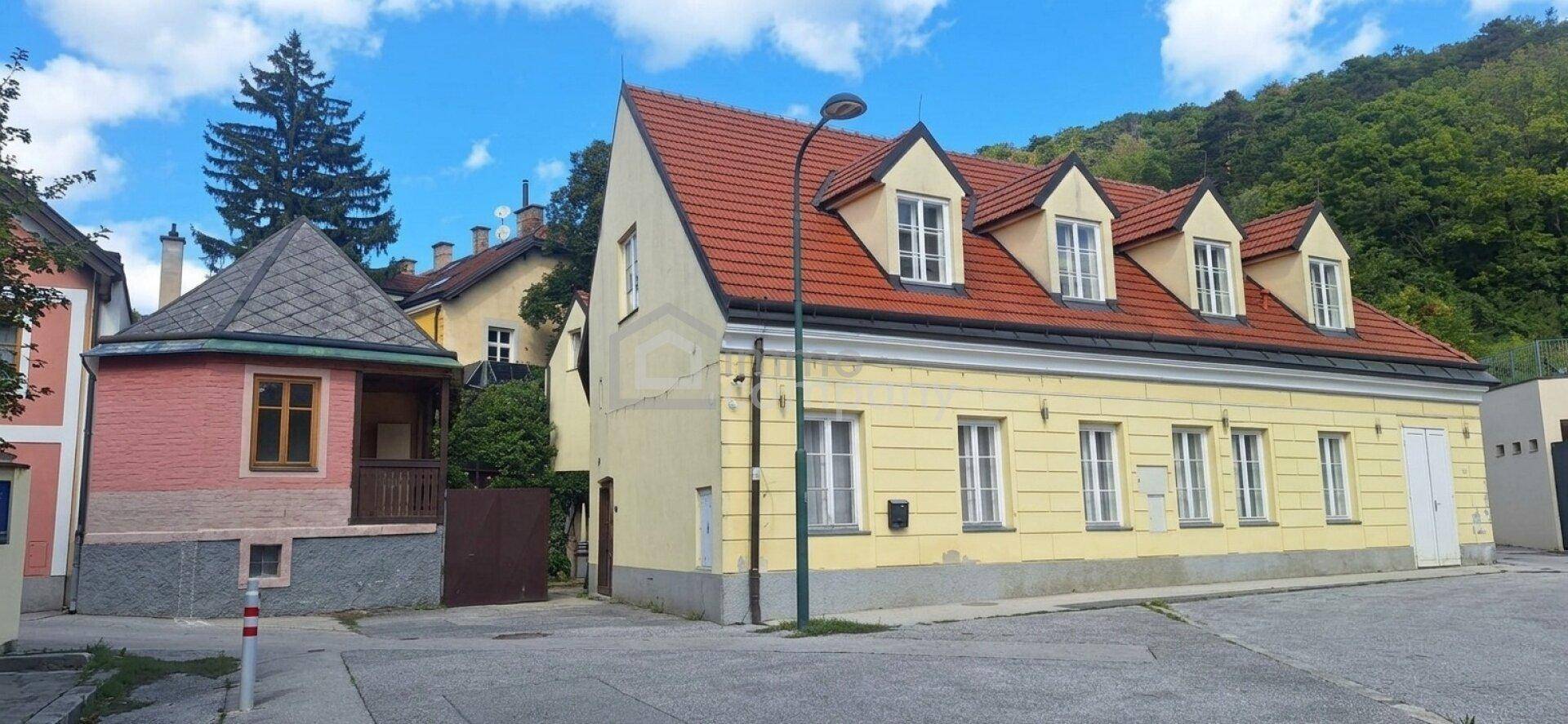 Traditionelles Gebäude mit gelber Fassade und rotem Ziegeldach, daneben ein kleineres rosa Gebäude an der Straße.