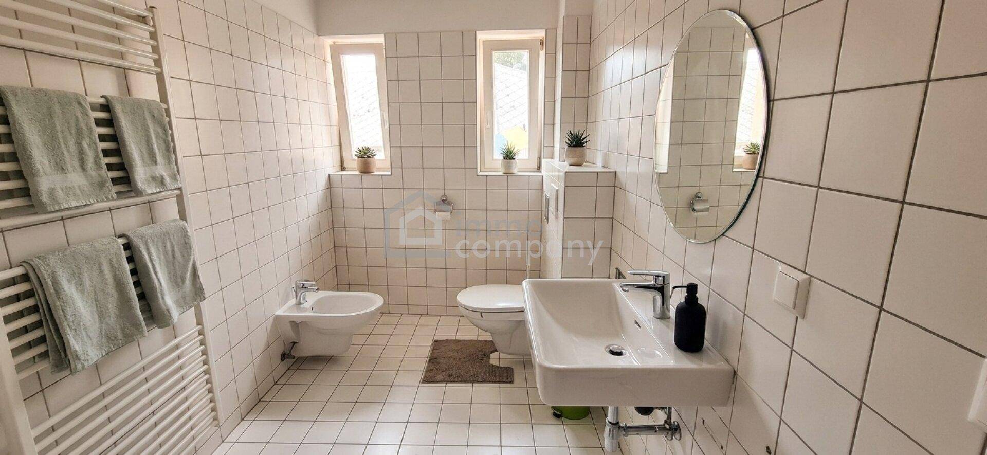 Helles Badezimmer mit weißen Fliesen, Waschbecken, Toilette, Bidet und beheiztem Handtuchhalter.