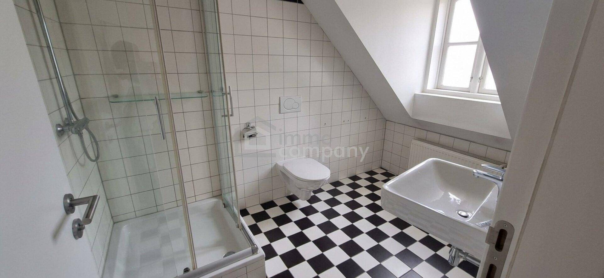 Badezimmer im Dachgeschoss mit Dusche, Toilette, Waschbecken und schwarz-weiß karierten Bodenfliesen.