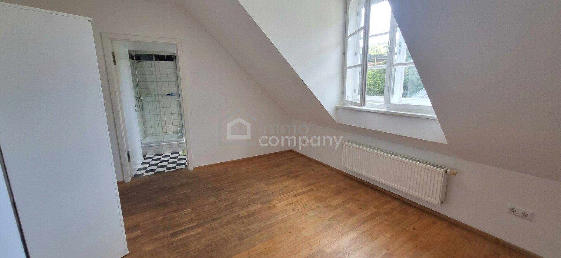 Helles Zimmer im Dachgeschoss mit Holzfußboden, Fenster und Zugang zu einem Badezimmer.